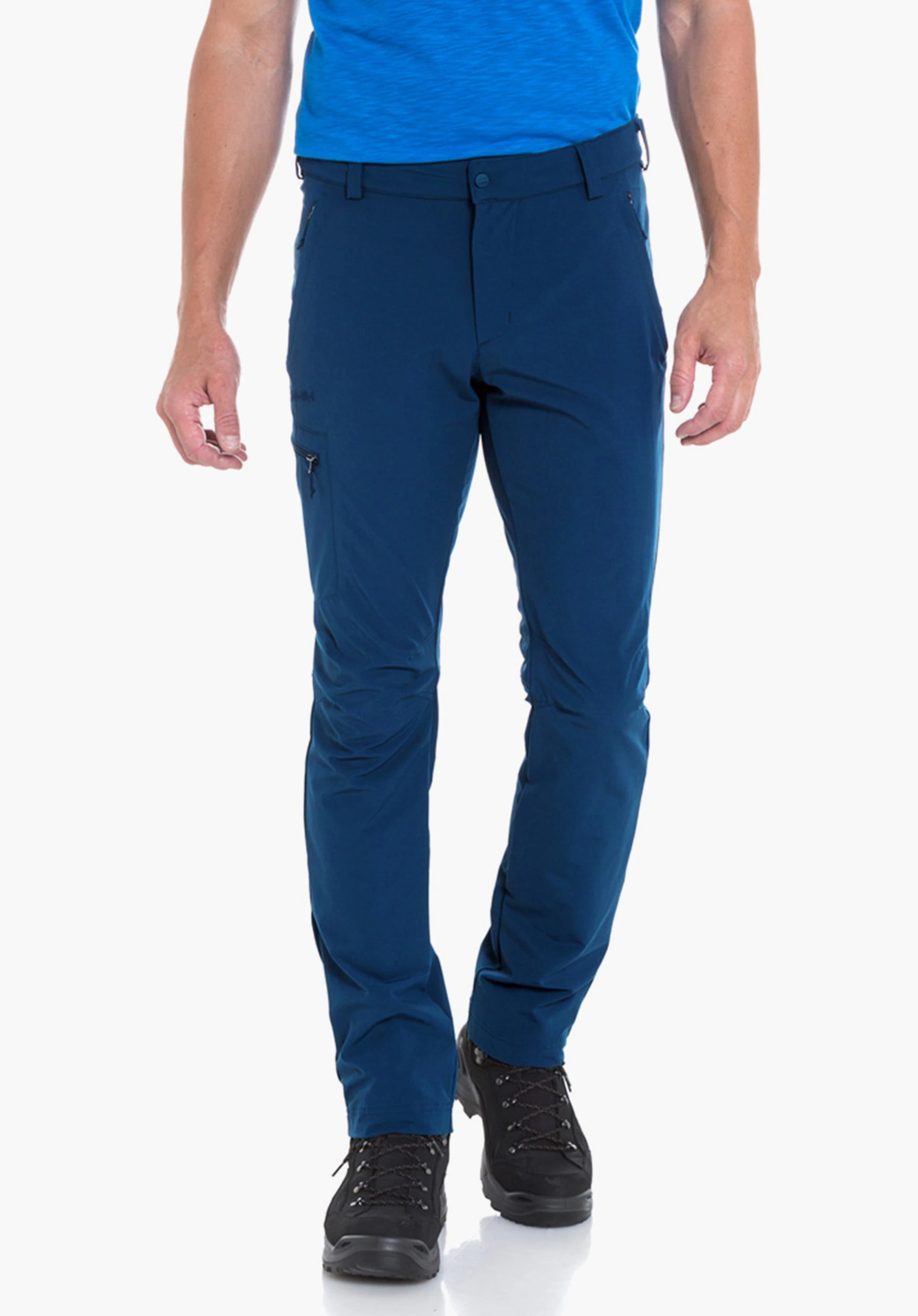 Pants Folkstone Homme porte un t-shirt bleu clair avec motif montagne et le texte « Schöffel », marche de manière décontractée; porte un pantalon bleu foncé et des chaussures de randonnée noires devant un fond blanc.