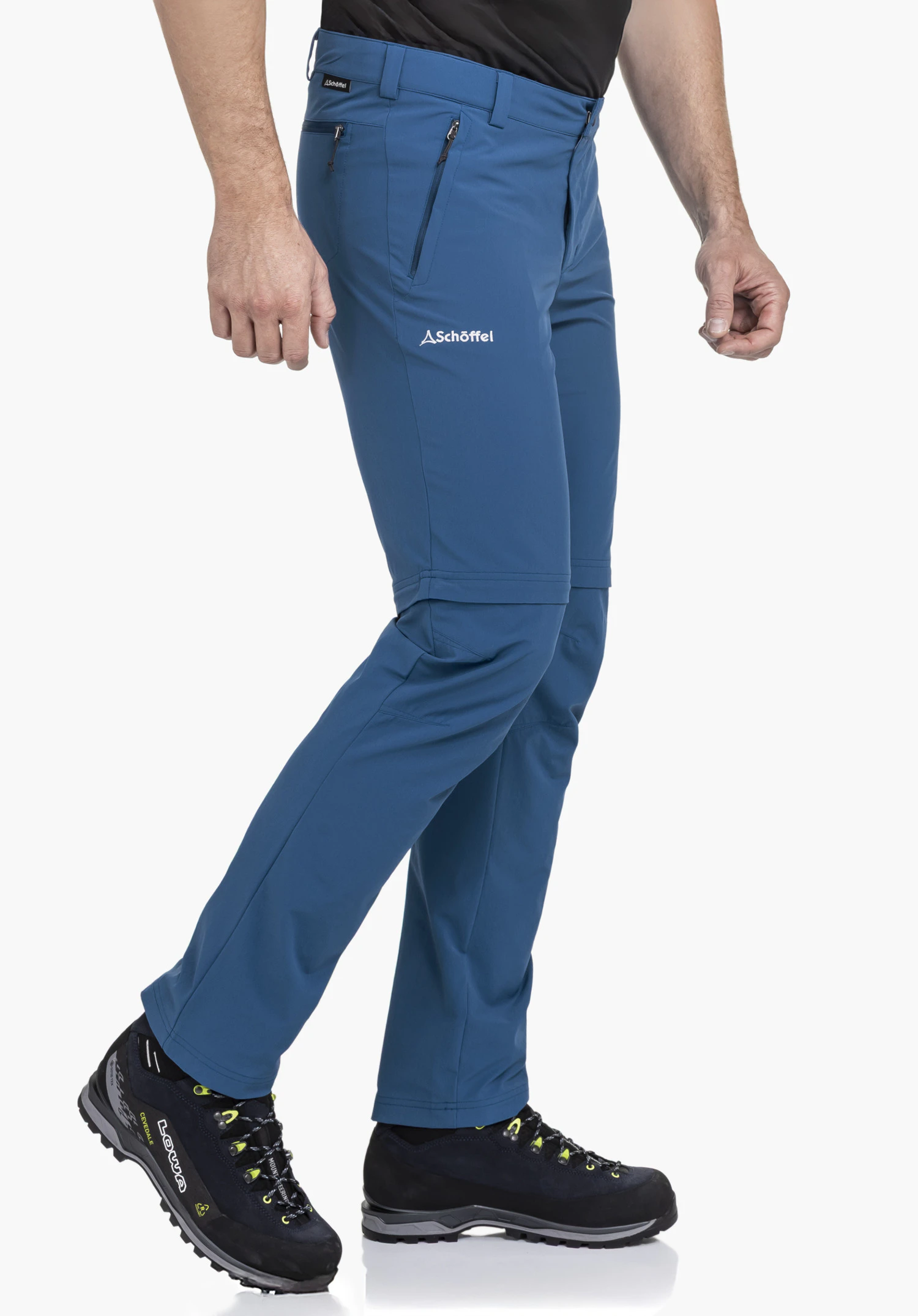 Pants Folkstone Zip Off. Een model staat zijwaarts in een wandelpose en draagt de blauwe broek. Het witte Schöffel-logo is goed zichtbaar op het linker bovenbeen. Het product wordt gepresenteerd tegen een neutrale achtergrond.