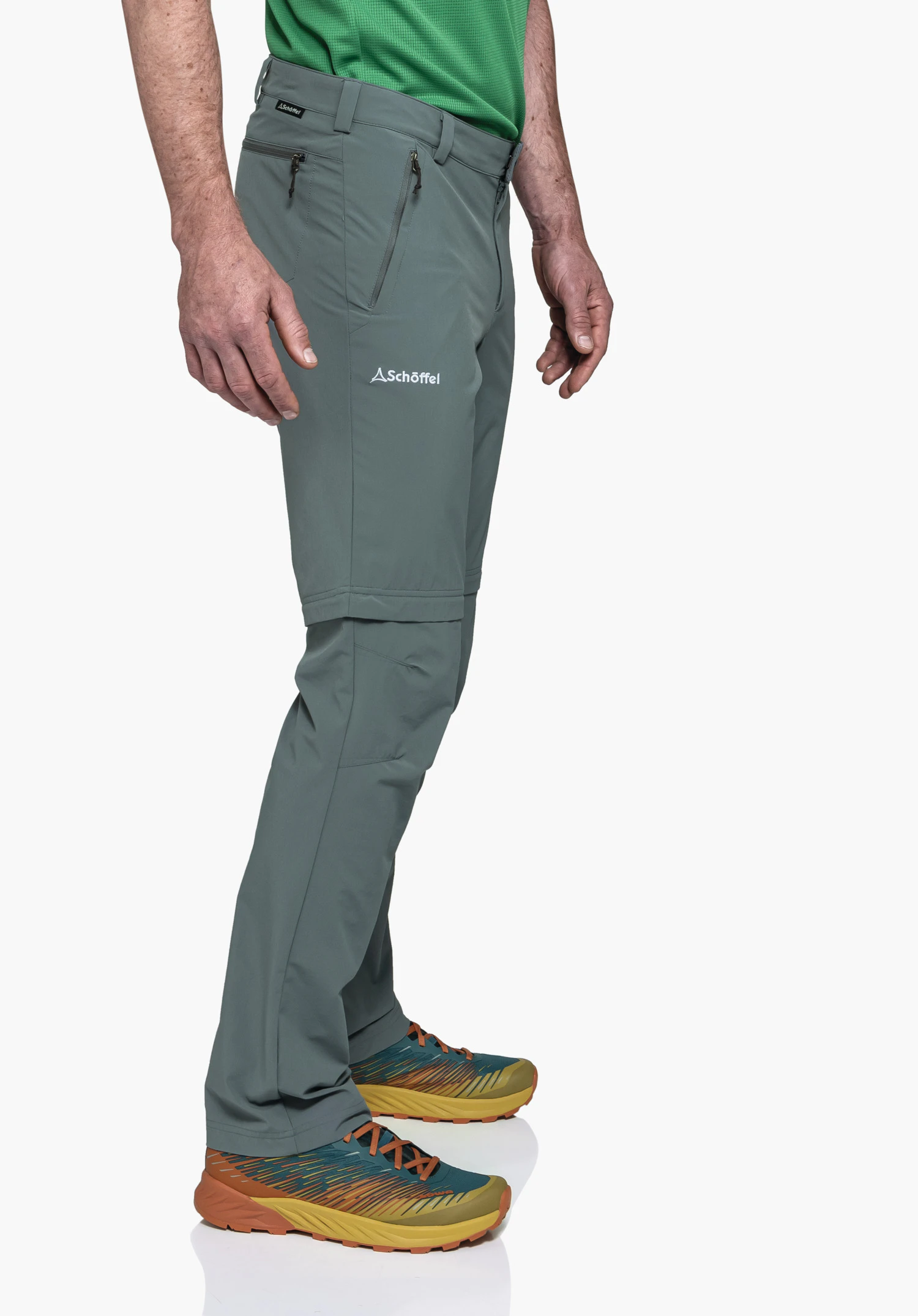 Pants Folkstone Zip Off gedragen door een model dat zijwaarts staat en de broek in de kleur grijs presenteert. Het Schöffel-logo is duidelijk zichtbaar op de linkerdij, evenals de rits voor de afritsfunctie op het been, gefotografeerd tegen een neutrale achtergrond.