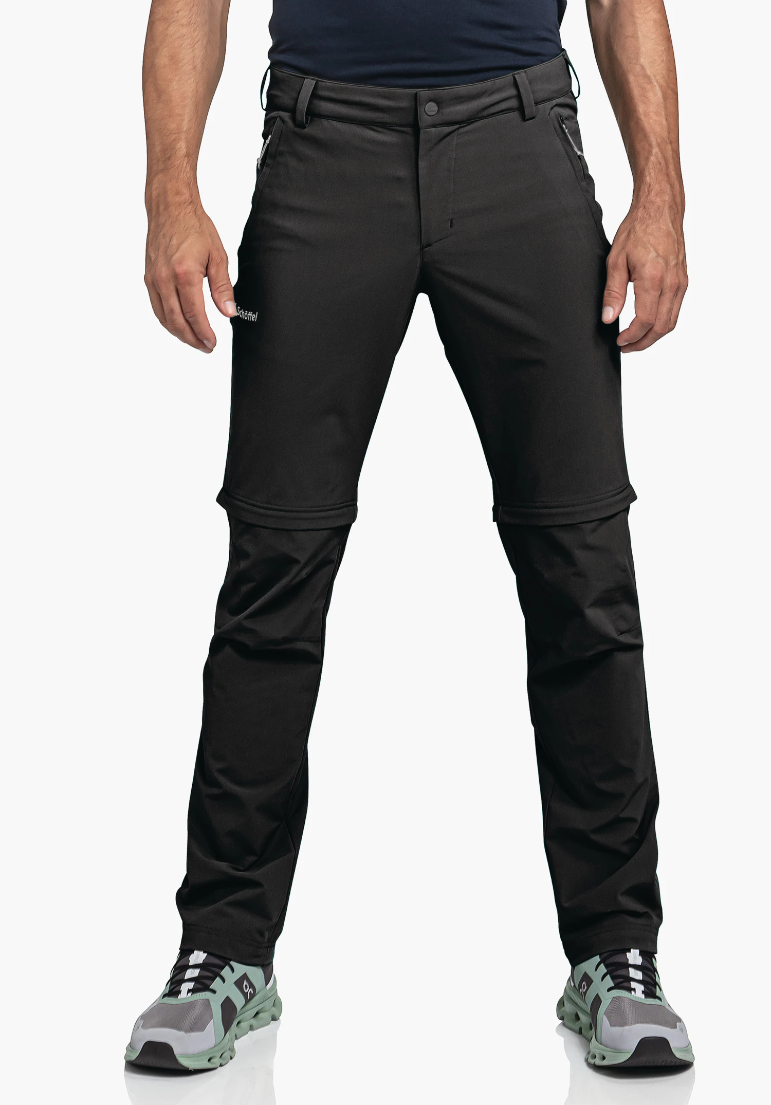 Pants Folkstone Zip Off en noir. Un modèle se tient de face et porte le pantalon avec des fermetures éclair visibles pour la fonction zip-off et le logo Schöffel sur la cuisse droite devant un fond neutre.