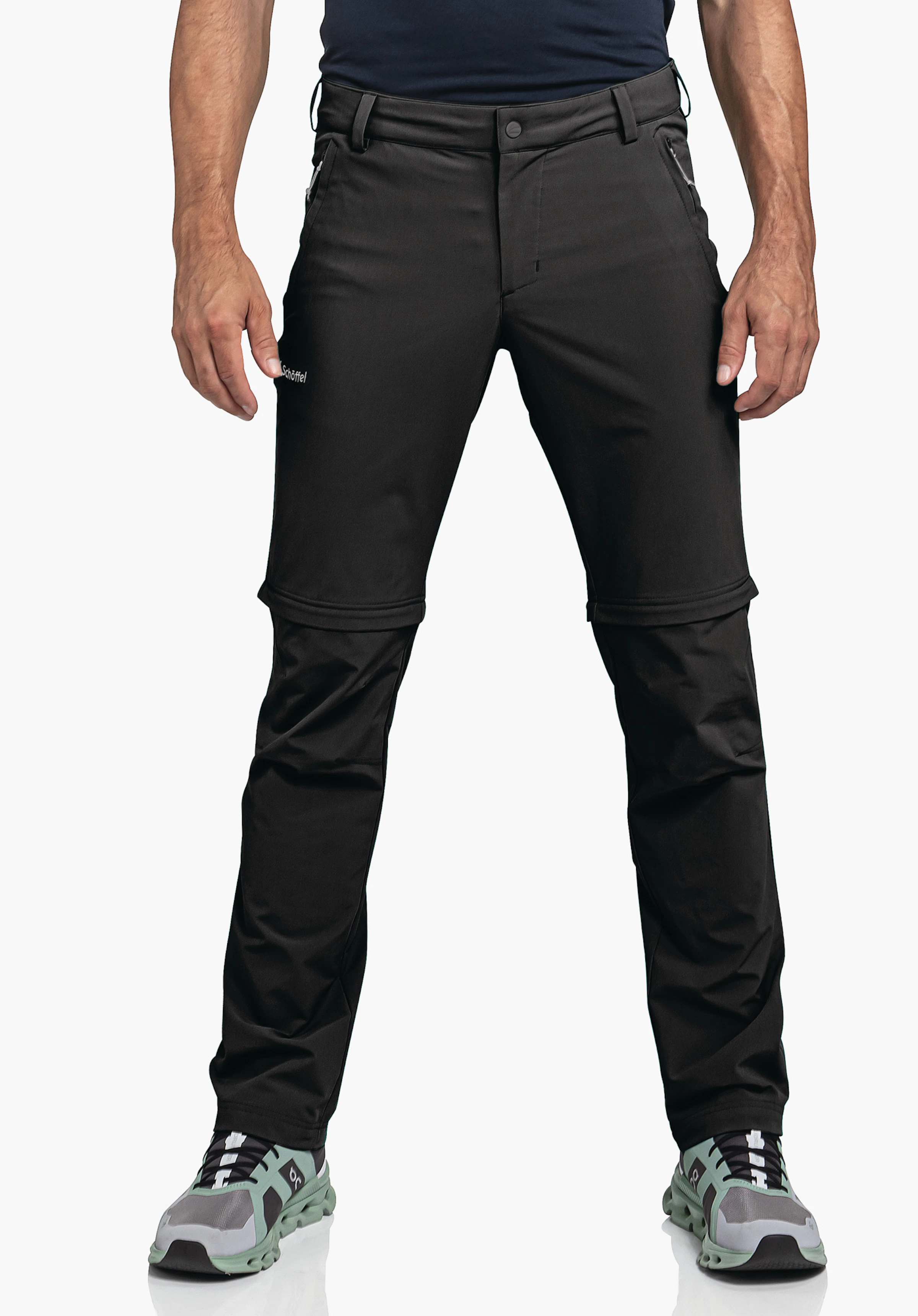 Pants Folkstone Zip Off en noir. Un modèle se tient de face et porte le pantalon avec des fermetures éclair visibles pour la fonction zip-off et le logo Schöffel sur la cuisse droite devant un fond neutre.