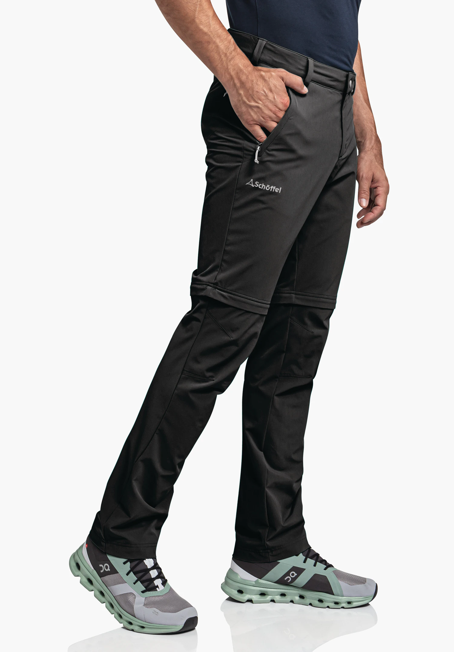 Pants Folkstone Zip Off sur un mannequin qui se tient de profil. Le pantalon en noir présente le Schöffel-Logo sur la cuisse et des fermetures éclair distinctes au-dessus du genou. Le modèle a une main nonchalamment glissée dans la poche avant, photographié devant un fond neutre.