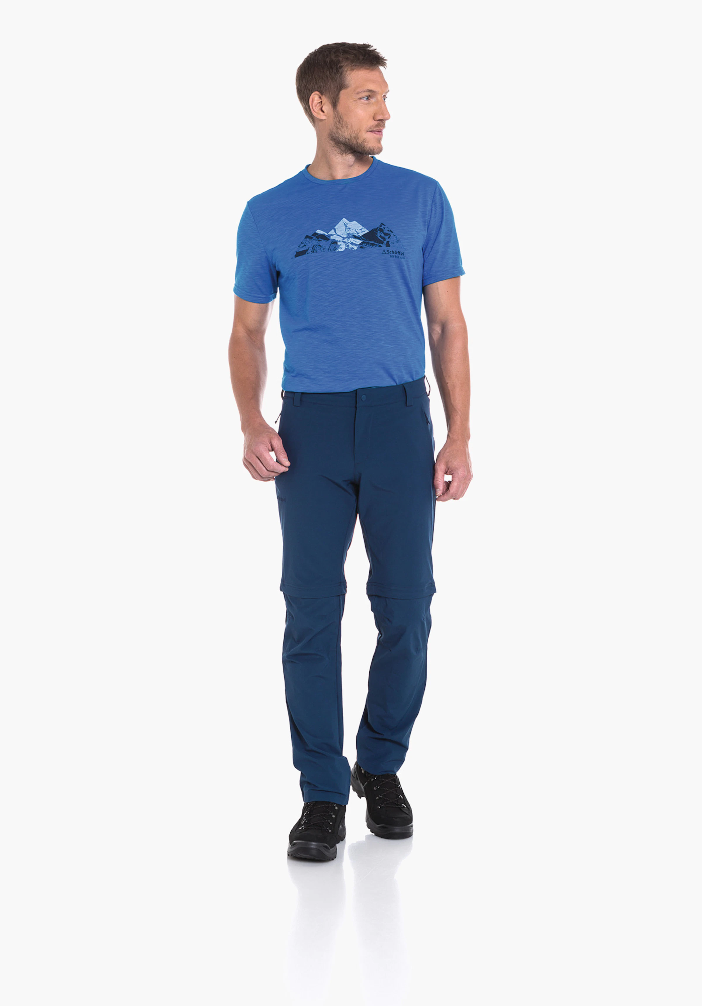 Pants Folkstone Zip Off Mann trägt hellblaues T‑Shirt mit Berggrafik und Schrift "Schöffel", steht und schaut nach rechts, trägt dunkelblaue Zip‑off‑Hose und schwarze Wanderschuhe, heller weißer Studiohintergrund.