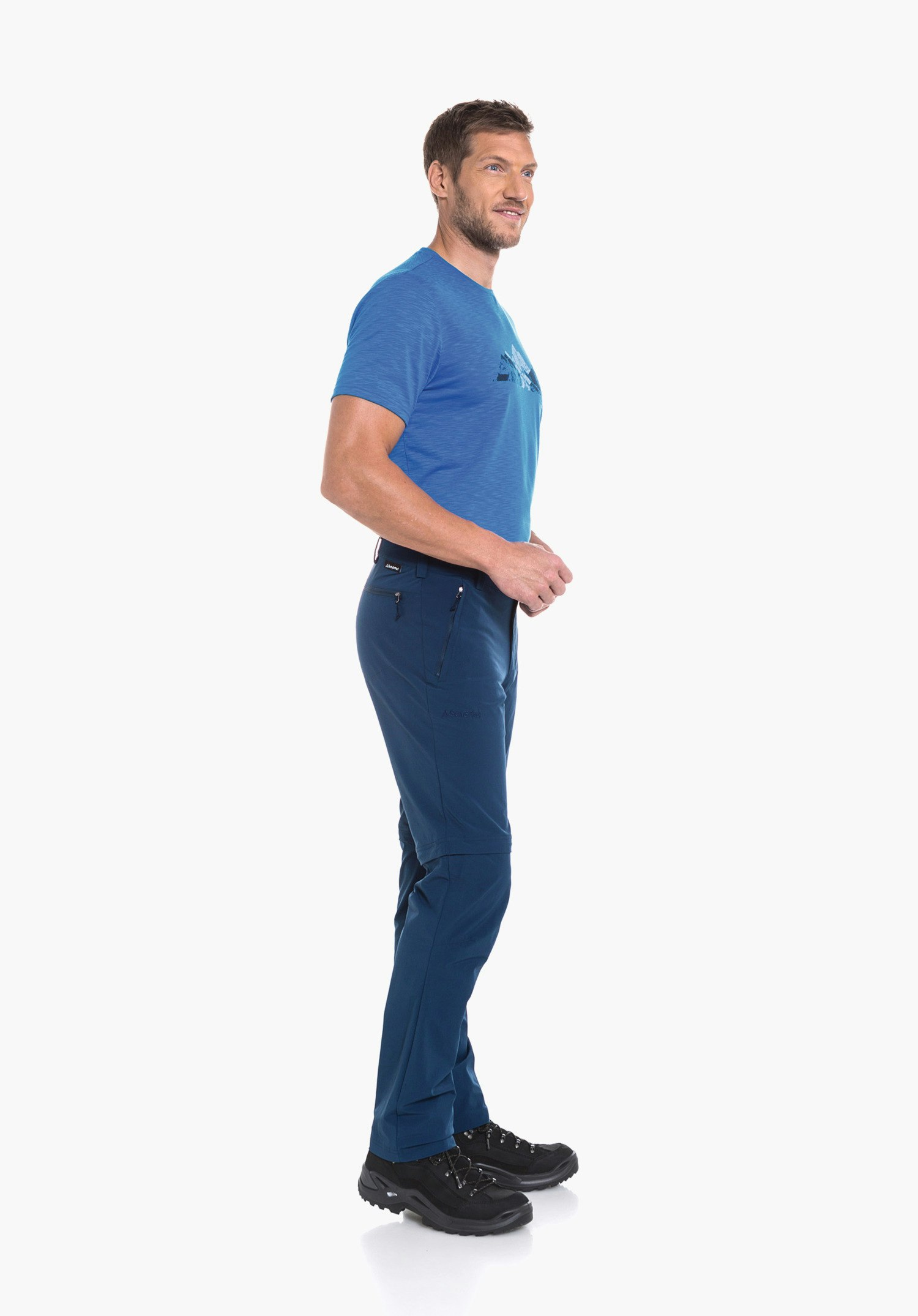 Pants Folkstone Zip Off man stands side-on wearing a light blue T-shirt and dark blue hiking trousers, looks to the right, hands on hips, black hiking boots; Schöffel logo on trousers.