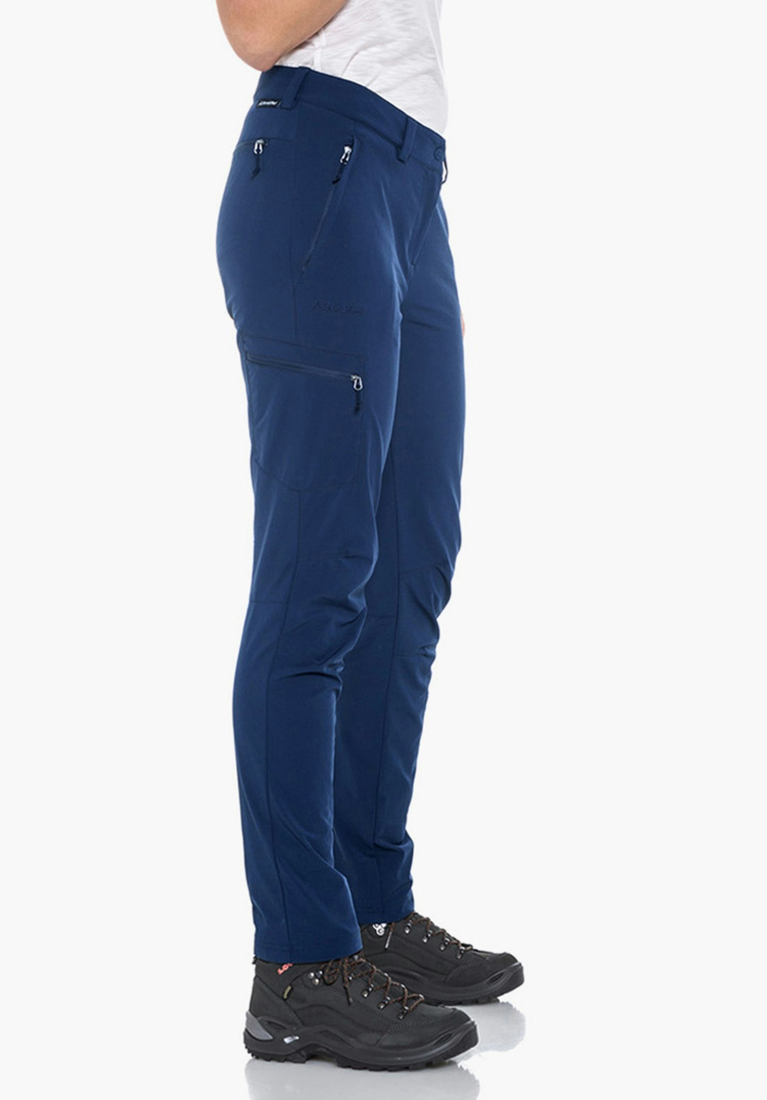Pants Ascona femme debout de profil, lève légèrement le bras droit; porte un t-shirt blanc, un pantalon bleu foncé et des chaussures de randonnée noires; cheveux bruns; fond clair neutre.