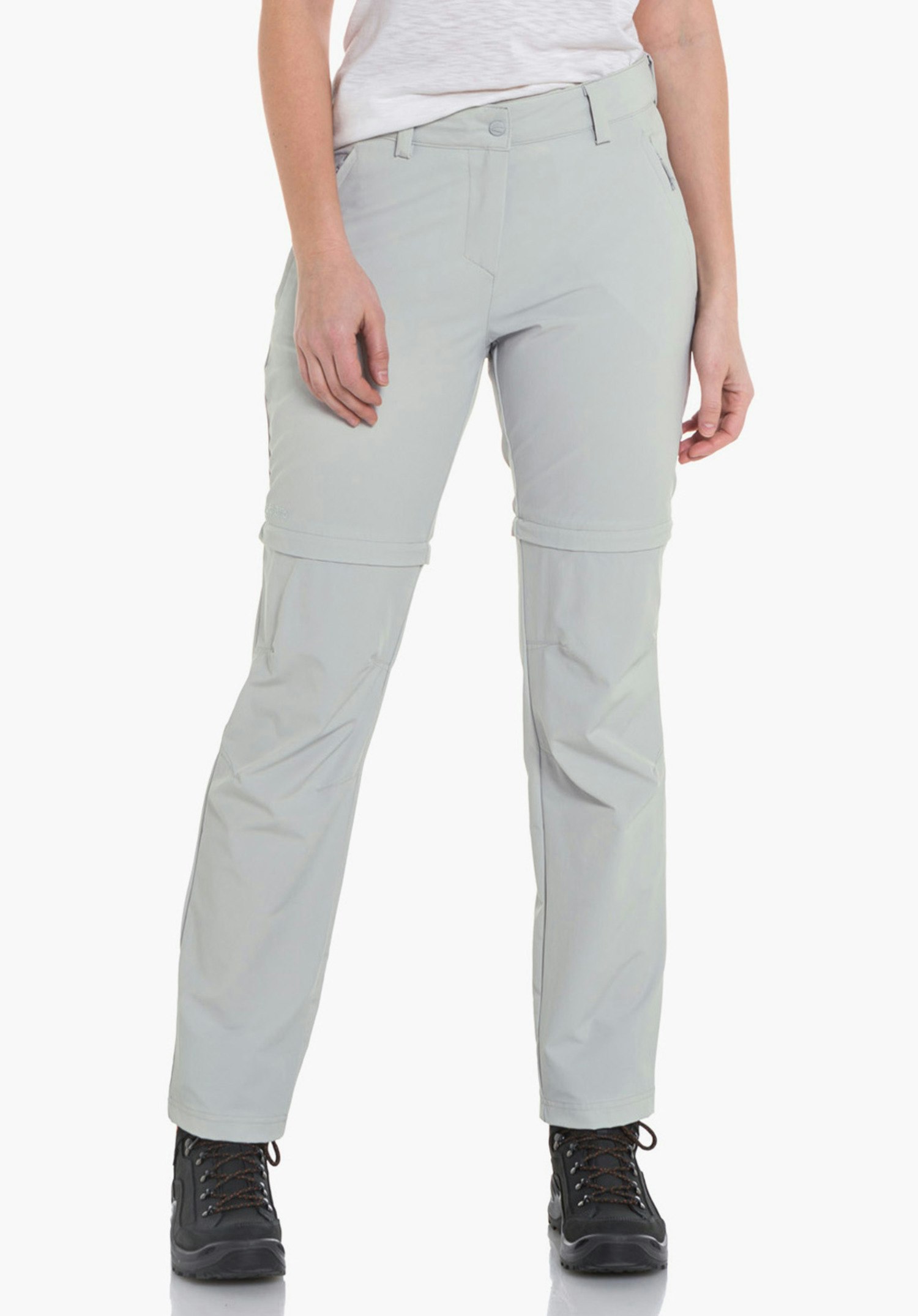 Pants Ascona Zip Off woman wears light grey zip-off trousers, standing relaxed and looking slightly to the side; combined with a white T-shirt and black hiking boots against a plain light background.