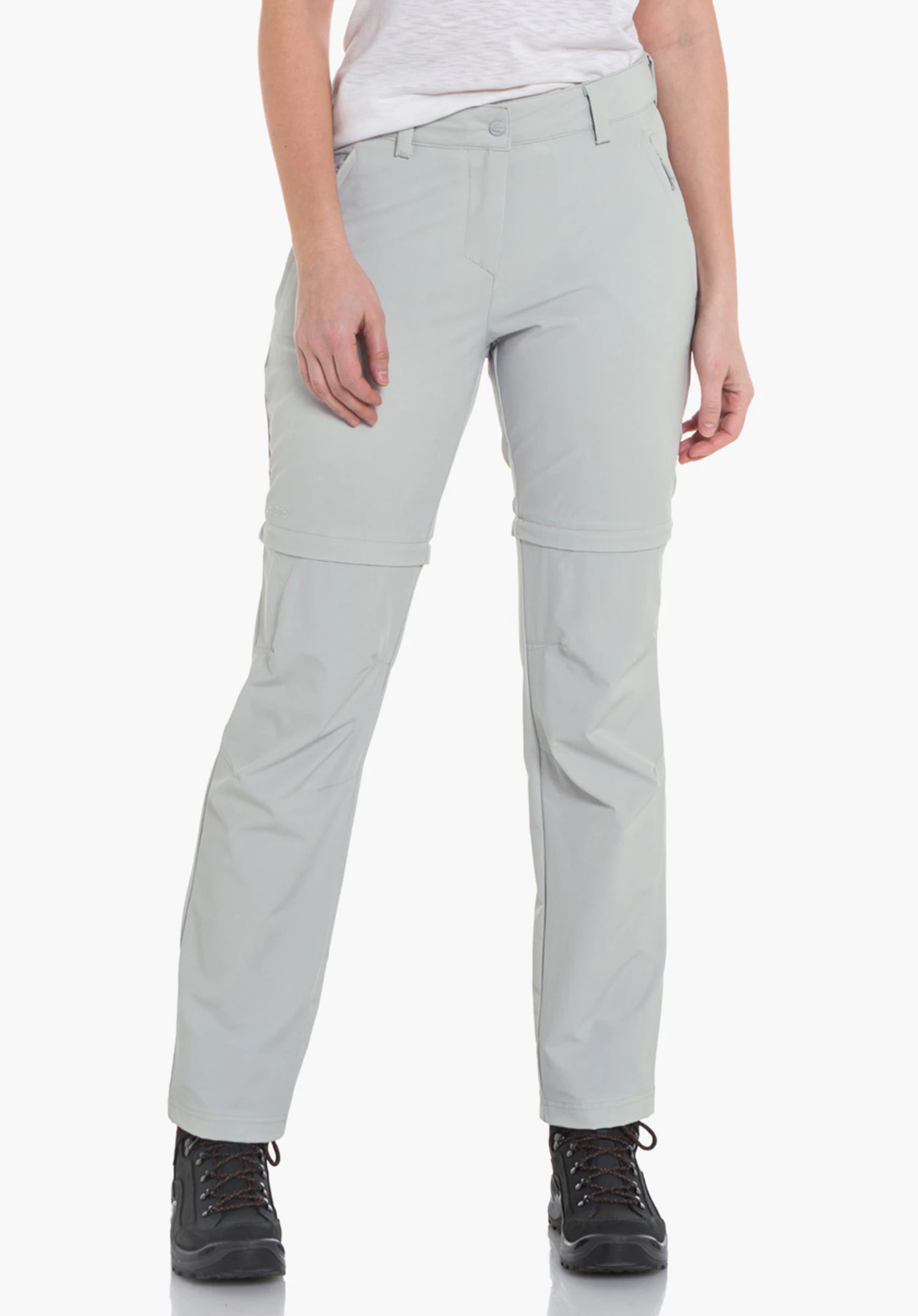 Pants Ascona Zip Off woman wears light grey zip-off trousers, standing relaxed and looking slightly to the side; combined with a white T-shirt and black hiking boots against a plain light background.