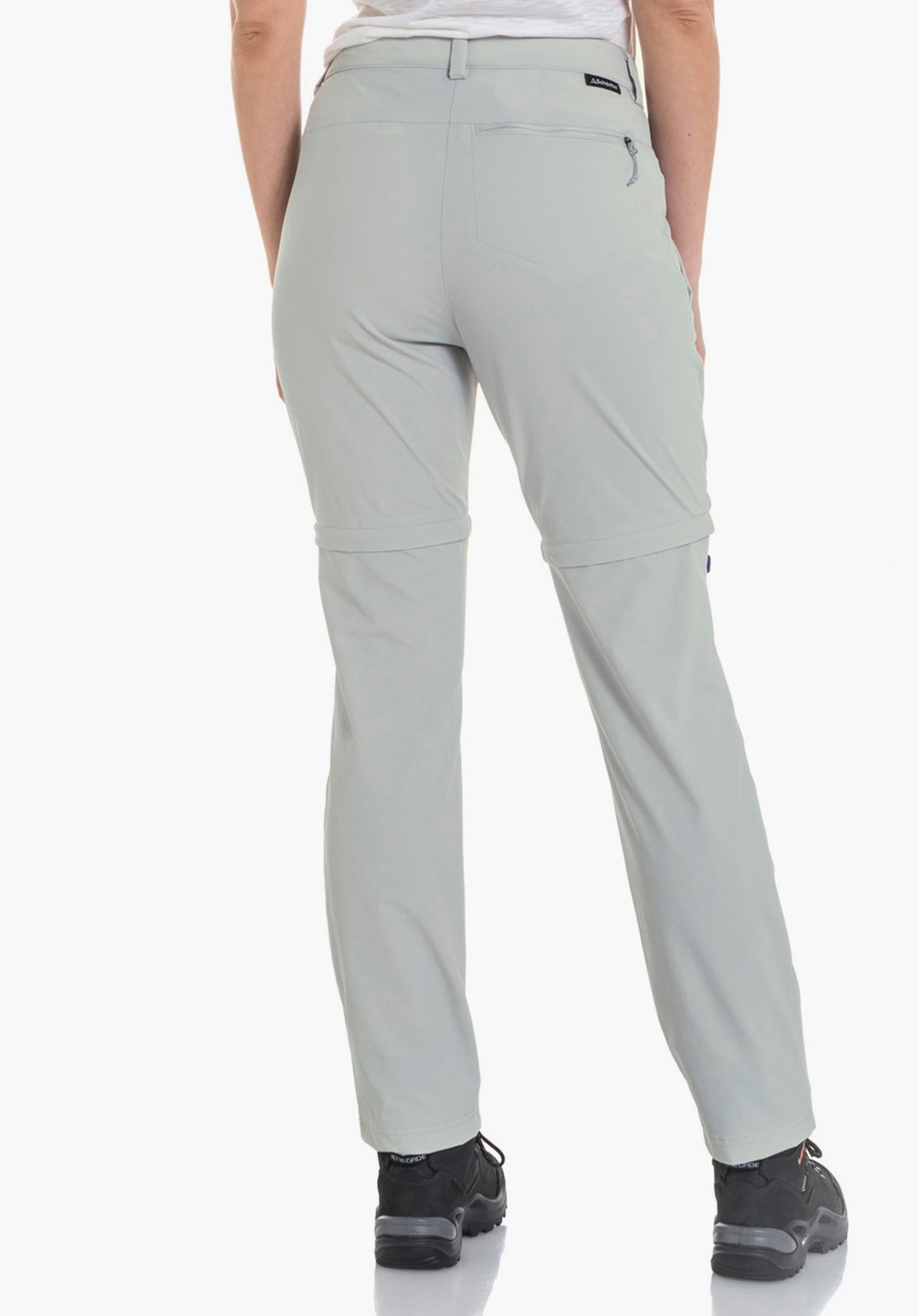 Pants Ascona Zip Off Damen-Zip-off-Hose, getragen von stehender Frau, Rückenansicht; sie trägt weißes T-Shirt; Hose hellgrau, kleine schwarze Schöffel-Logo am rechten Gesäß; helle Studioumgebung.