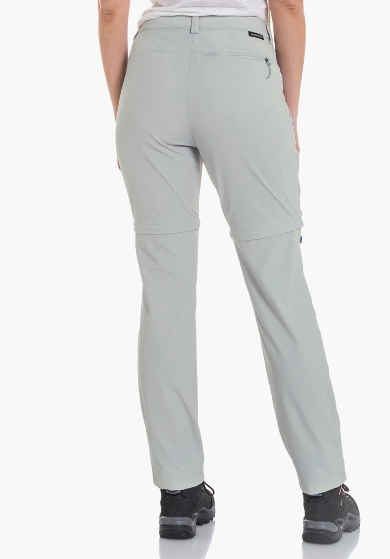 Pants Ascona Zip Off Damen-Zip-off-Hose, getragen von stehender Frau, Rückenansicht; sie trägt weißes T-Shirt; Hose hellgrau, kleine schwarze Schöffel-Logo am rechten Gesäß; helle Studioumgebung.