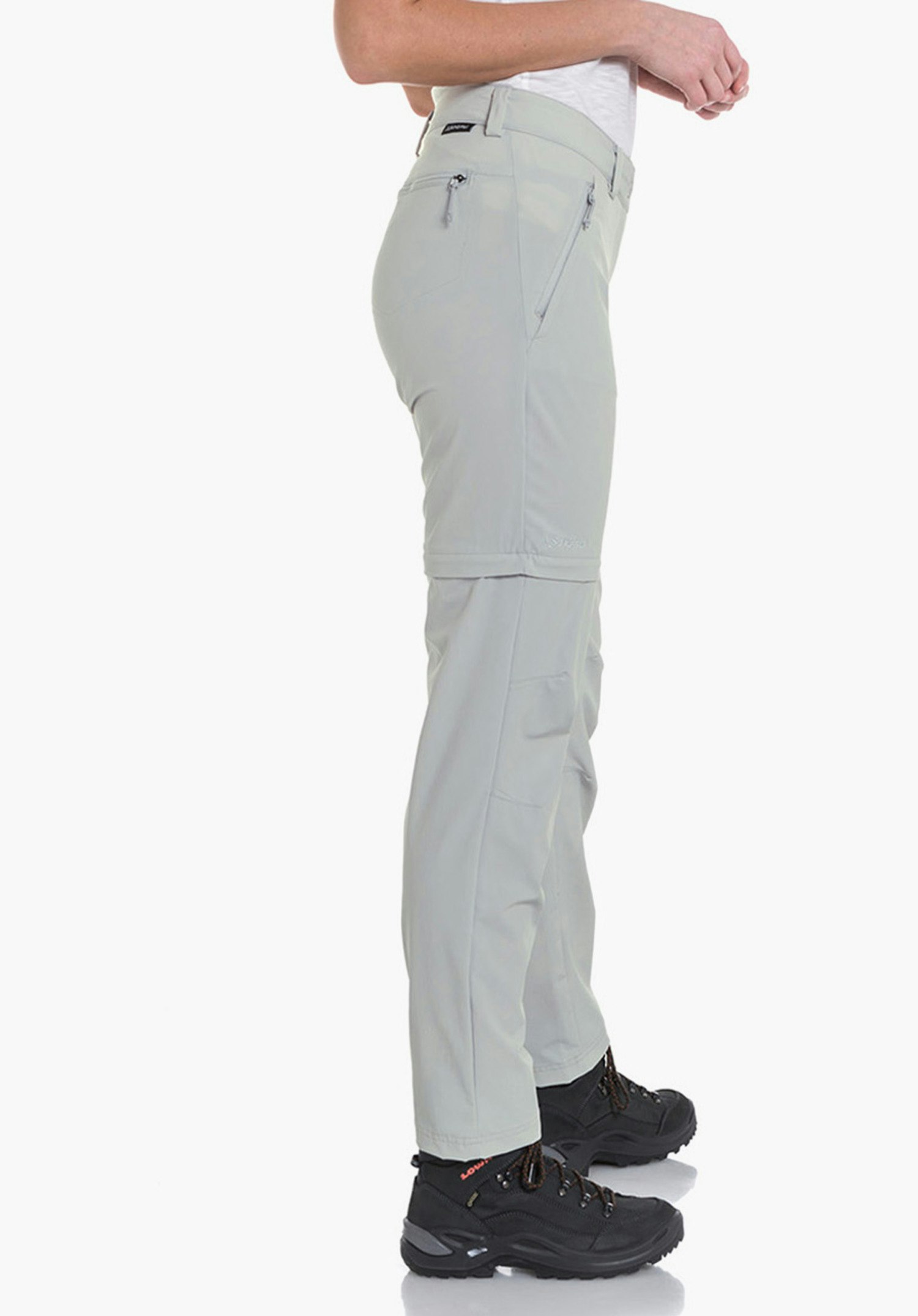Pants Ascona Zip Off Frau trägt hellgraue Zip-off Wanderhose, steht seitlich mit Händen an der Hüfte und blickt über die Schulter; dazu weißes T‑Shirt und schwarze Wanderschuhe, weißer Studiohintergrund.