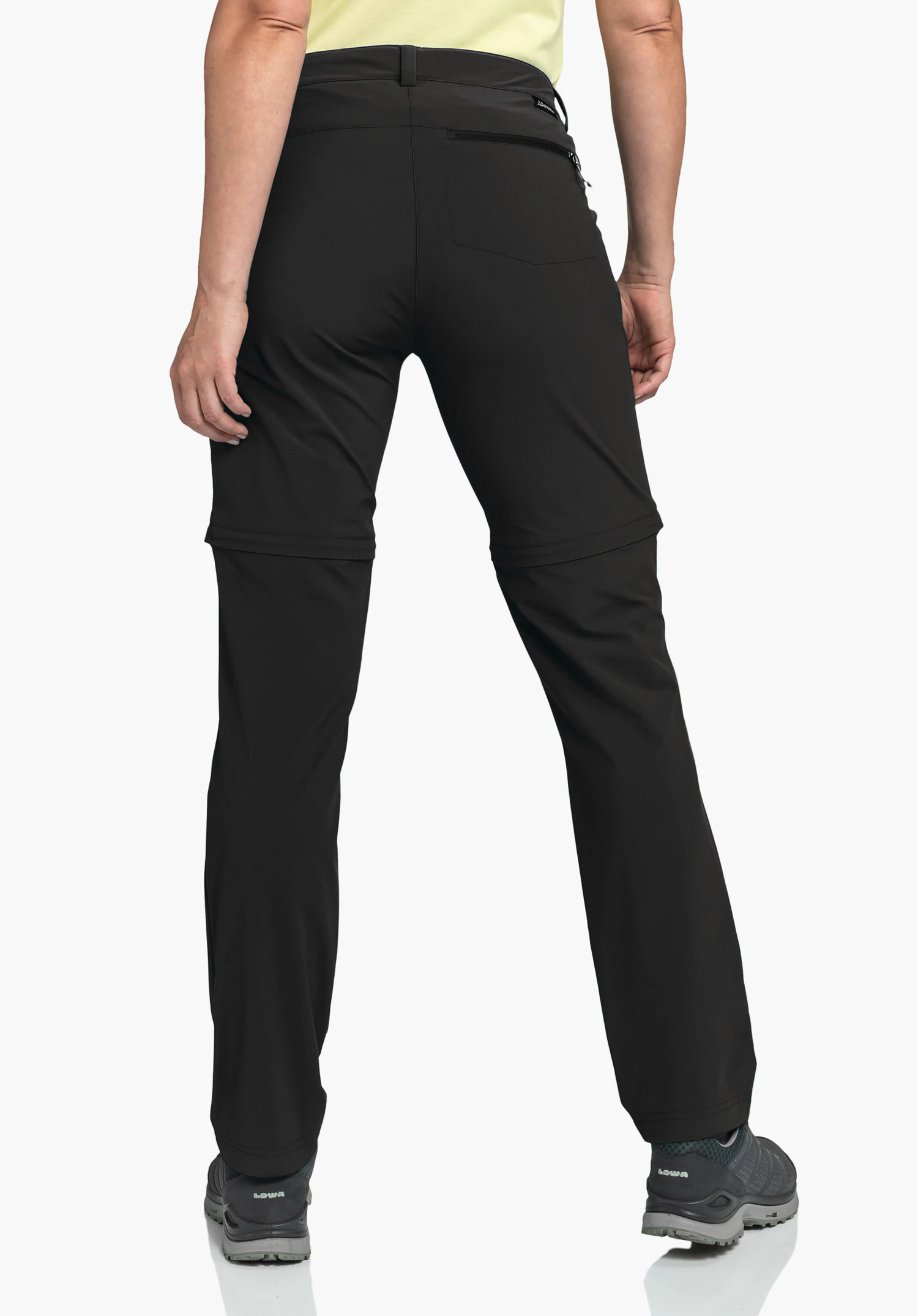 Pants Ascona Zip Off on a model in a back view. The black trousers show the zip-off zips on the legs and a right back pocket with a zip. A small Schöffel logo is visible on the side of the waistband. The model stands in front of a neutral background.