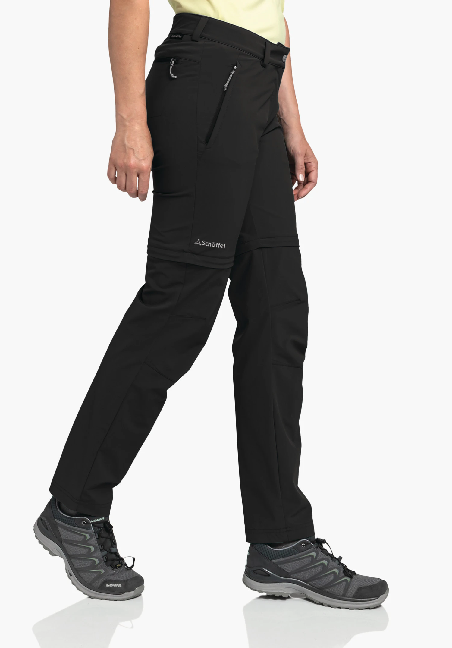 Pants Ascona Zip Off worn by a model standing sideways in a walking pose. The black trousers feature a side zip pocket and the Schöffel-Logo on the thigh just above the zip-off seam against a neutral background.