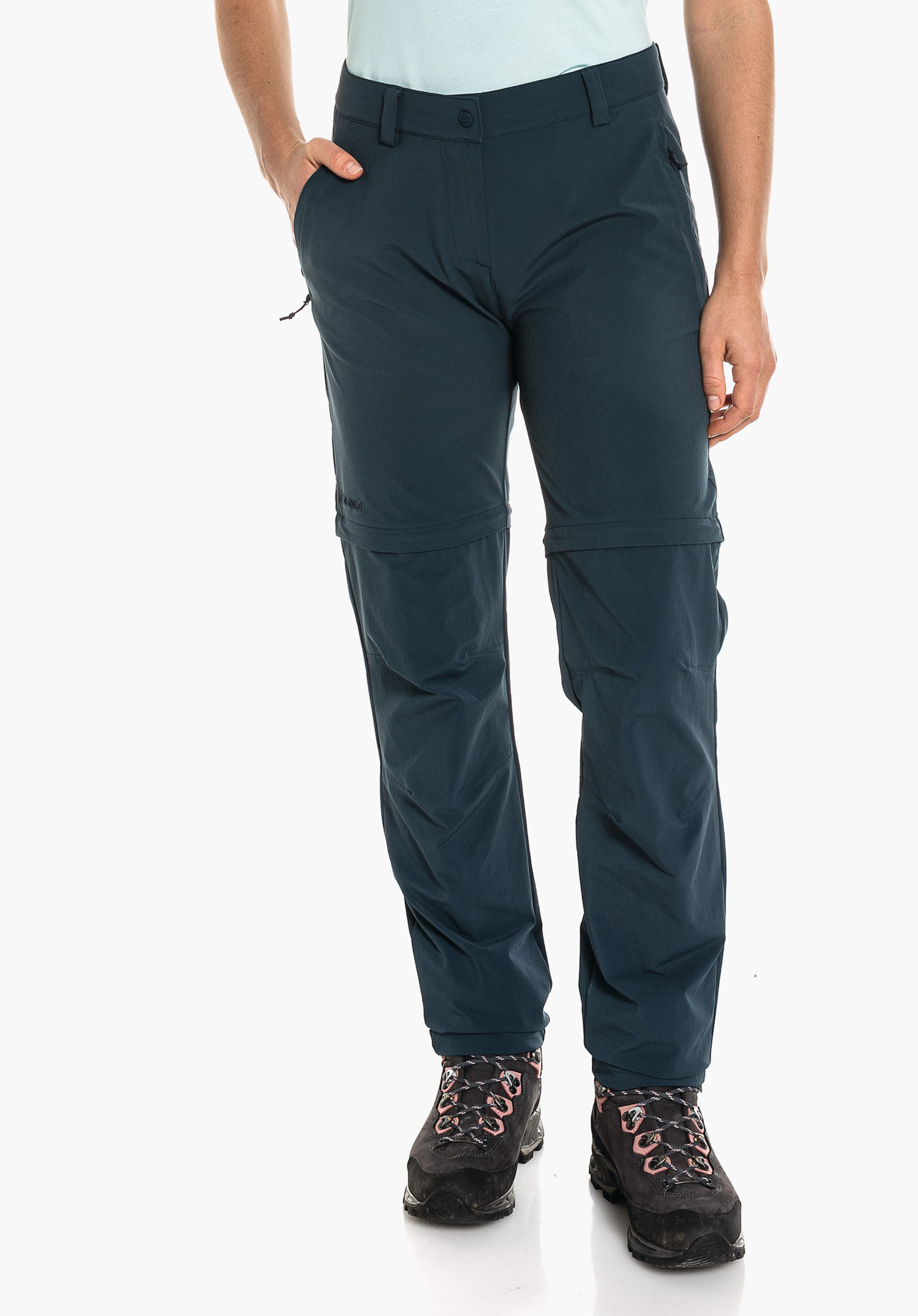 Pants Ascona Zip Off Dunkelblaue Zip-off-Hose, getragen von stehender Person; eine Hand in Tasche, hellblaues Oberteil, schwarze Wanderschuhe, neutraler weißer Studiohintergrund.