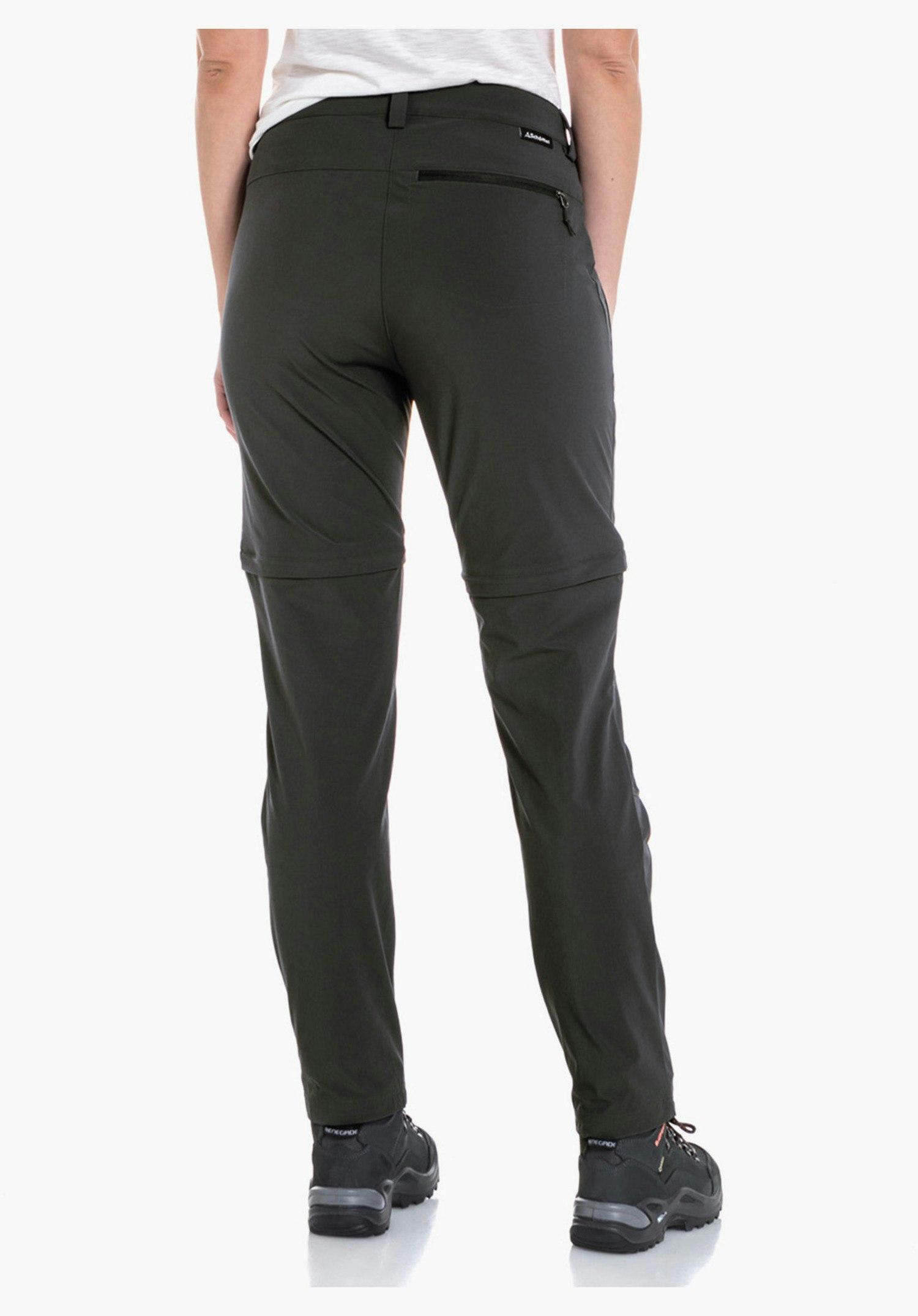 Pants Ascona Zip Off Damenhose dunkelgrün, getragen von Frau; sie steht mit dem Rücken zur Kamera, schaut nach rechts, Hände in den Taschen; weiße Umgebung, schwarze Schuhe.