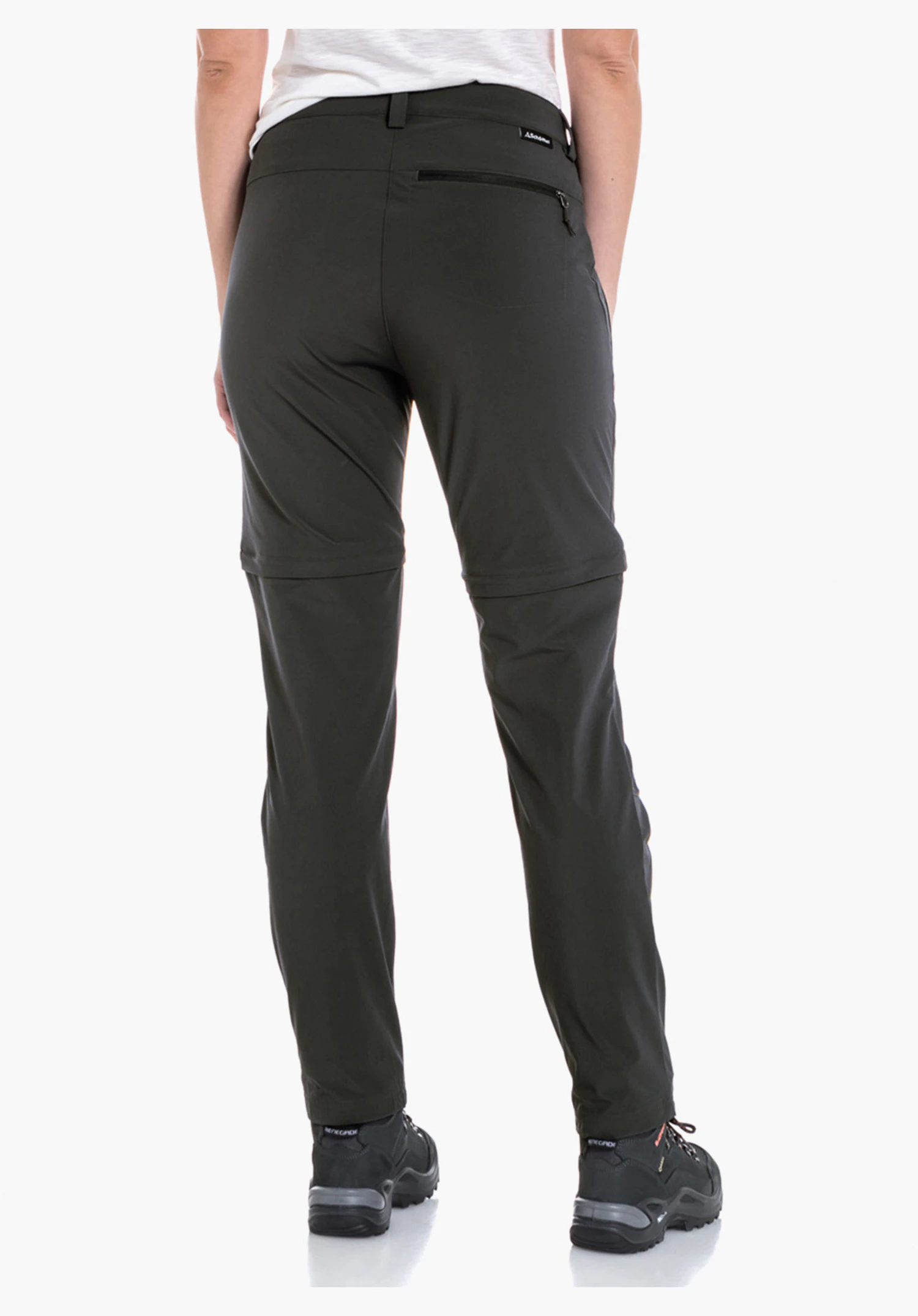 Pants Ascona Zip Off Damenhose dunkelgrün, getragen von Frau; sie steht mit dem Rücken zur Kamera, schaut nach rechts, Hände in den Taschen; weiße Umgebung, schwarze Schuhe.