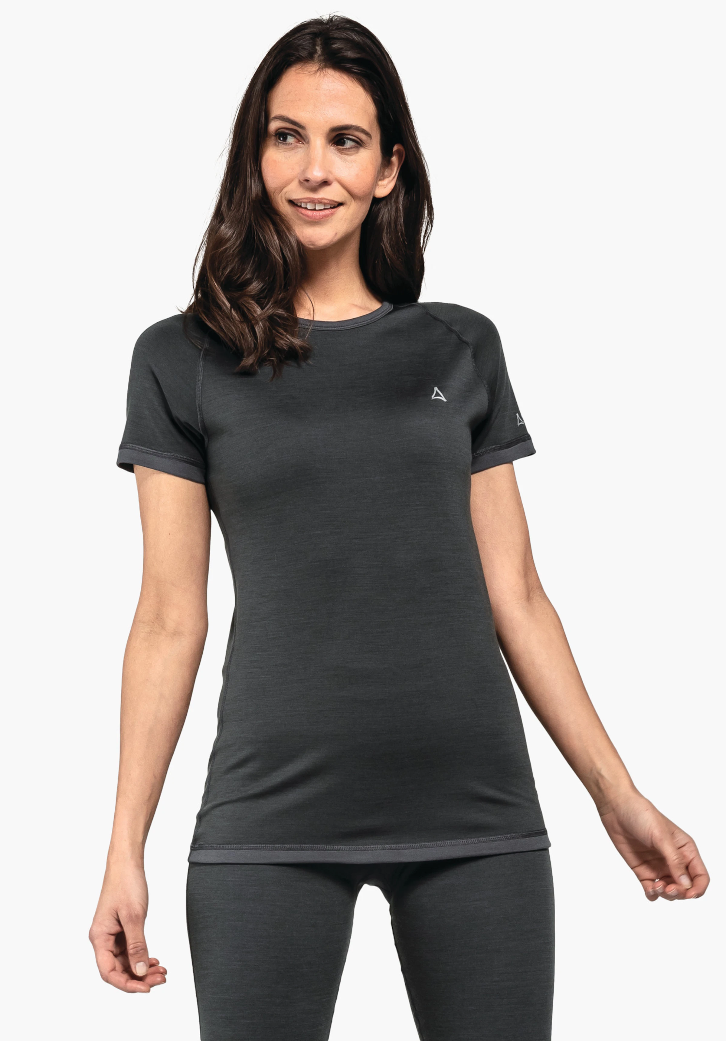 Merino Sport Shirt 1/2 Arm W Woman wears a fitted short-sleeve shirt with Schöffel logo; stands relaxed, looks to the side and smiles slightly, arms loosely lowered against a plain background.