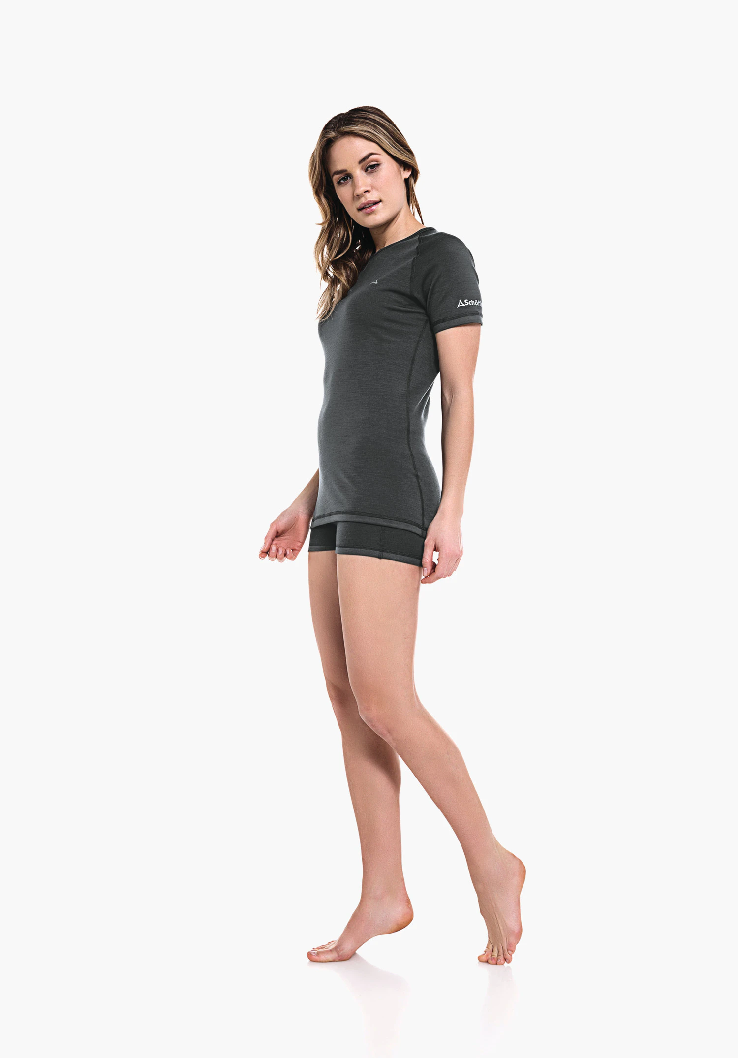 Merino Sport Shirt 1/2 Arm W woman in a dark grey sports shirt and shorts stands slightly side-on on tiptoes, arms relaxed; white background. On sleeve Schöffel logo and lettering "Schöffel".