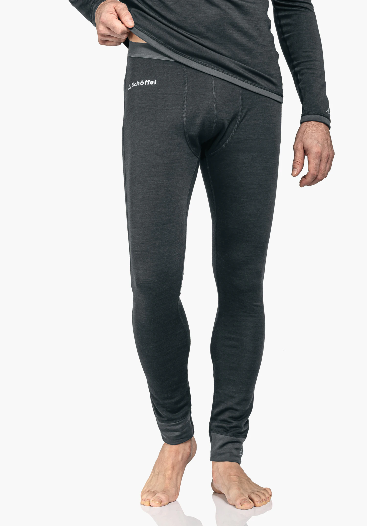 Merino Sport Pants long M Dark grey long thermal underwear; man slightly lifts top, stands barefoot against a white background; “Schöffel” lettering and Schöffel logo visible on the garment.