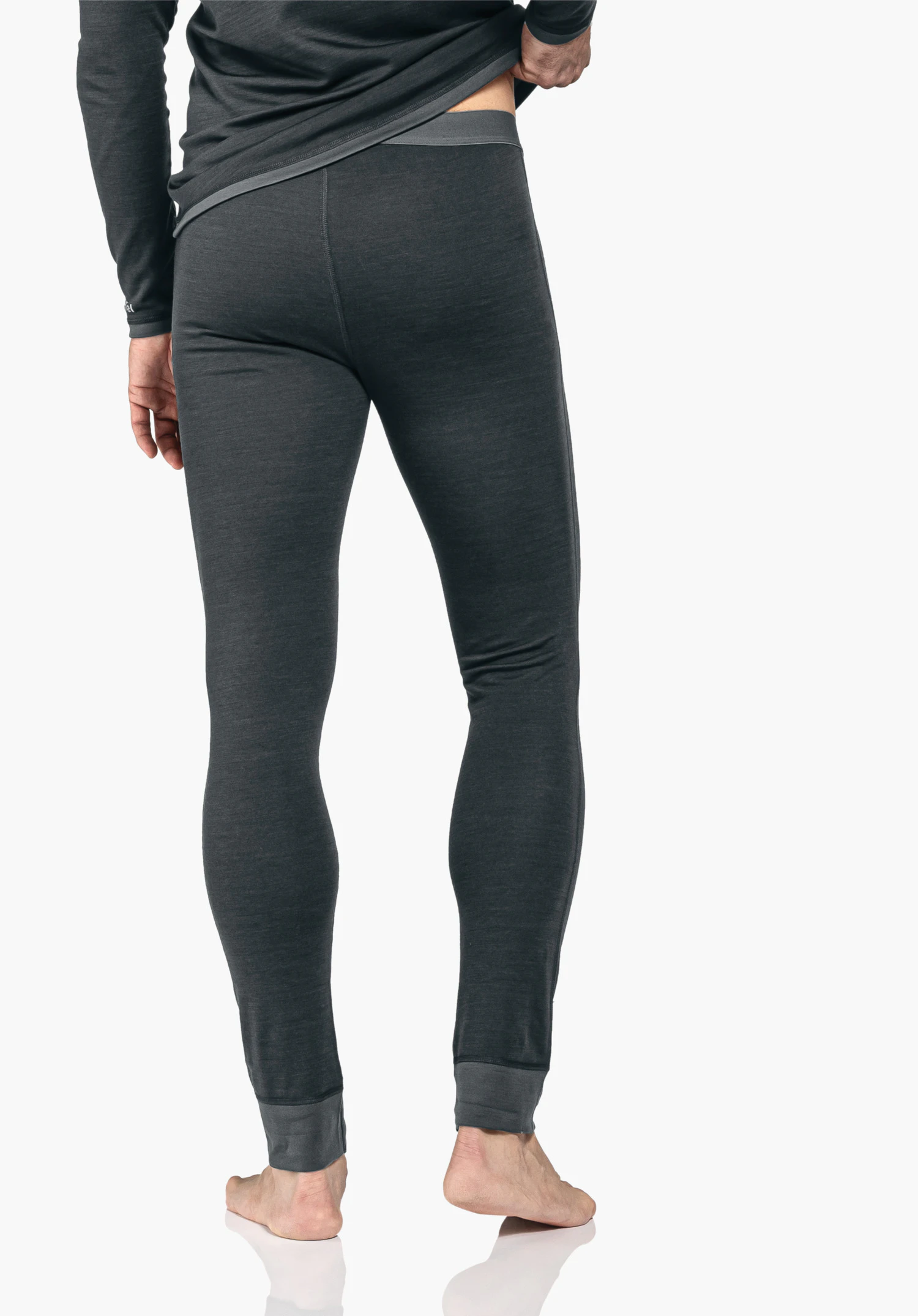 Merino Sport Pants long M black long underwear worn from the back; person slightly lifts the shirt and stands barefoot against a light plain background.