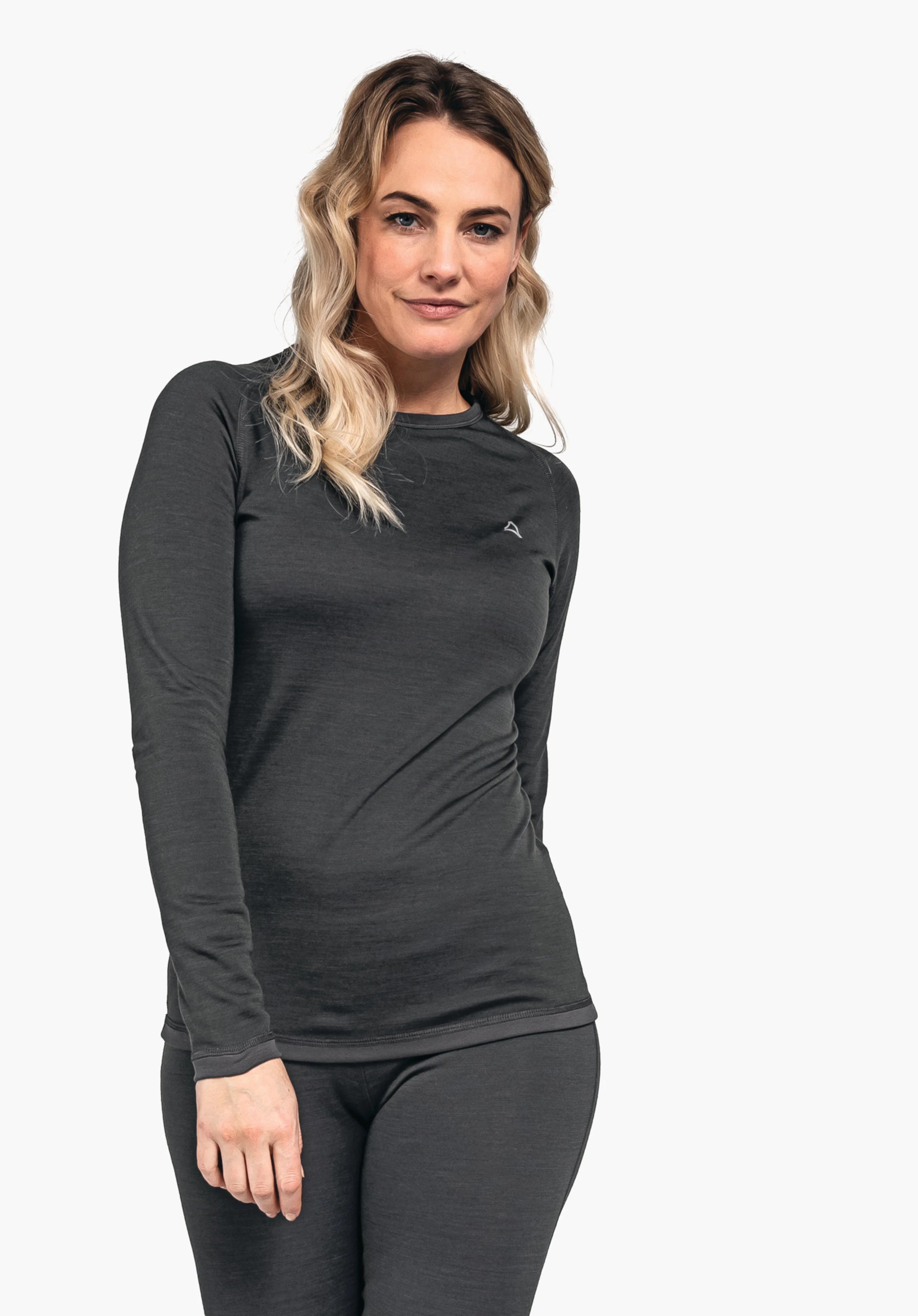 Merino Sport Shirt 1/1 Arm W Frau trägt dunkelgraues Langarmshirt und Leggings mit Schöffel-Logo, steht leicht nach vorn geneigt, lächelt dezent, rechte Hand entspannt vor dem Körper; heller neutraler Hintergrund.