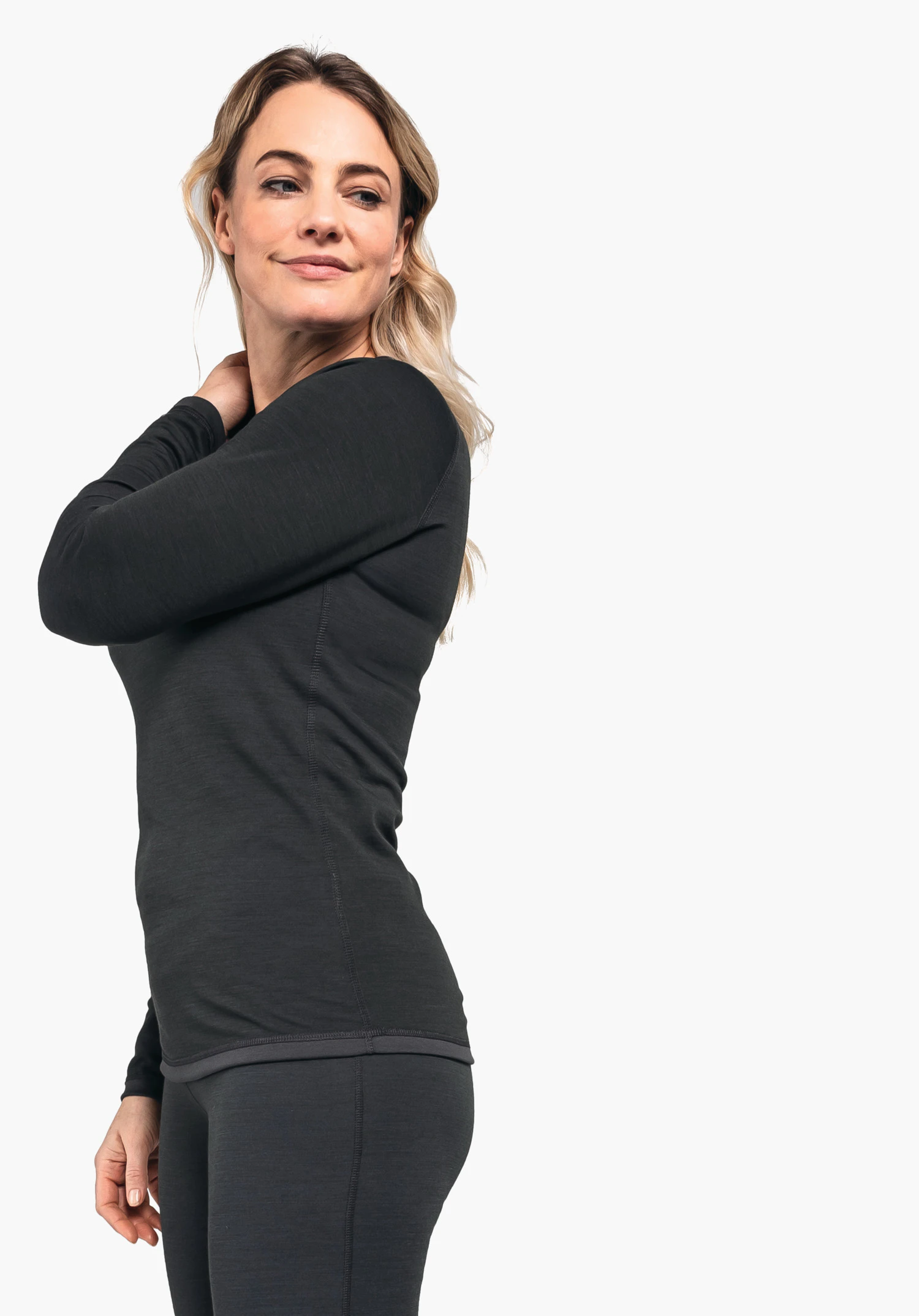 Merino Sport Shirt 1/1 Arm W Frau in dunkelblauem langärmligem Oberteil und dunkelblauen Leggings dreht den Oberkörper leicht nach rechts und fasst den Nacken; weißer Hintergrund.