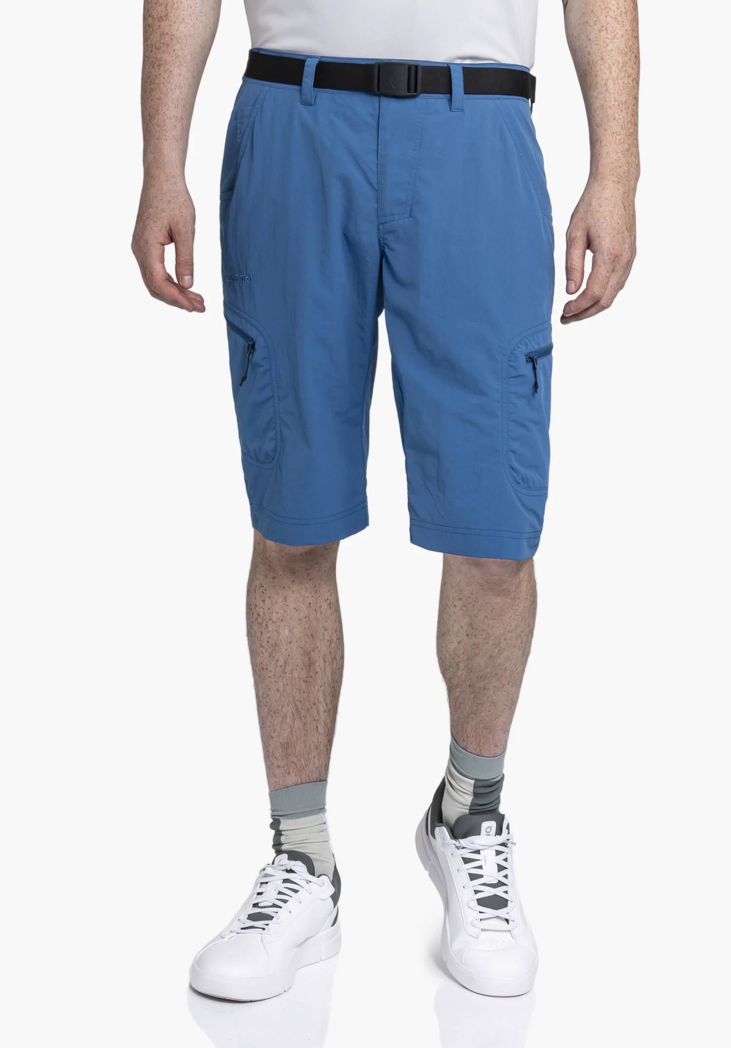 Shorts Silvaplana2 an einem Model in der Frontalansicht. Das Model steht frontal und trägt die blaue kurze Hose mit Gürtel und sichtbarem Schöffel-Logo auf dem rechten Bein vor einem neutralen Hintergrund.