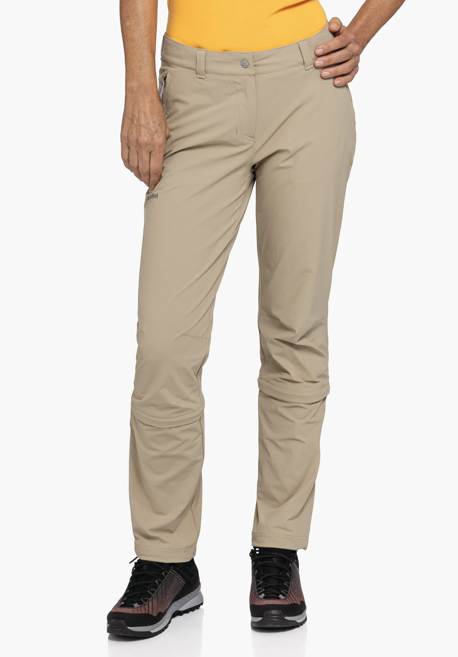 Pants Engadin1 Zip Off. Ein Model steht in der Frontalansicht und präsentiert die beige Hose mit sichtbaren Zip-Off-Reißverschlüssen über den Knien und dem Schöffel-Logo auf dem rechten Oberschenkel. Die Person posiert mit einer Hand an der Hüfte vor einem neutralen Hintergrund und trägt dazu dunkle Wanderschuhe.