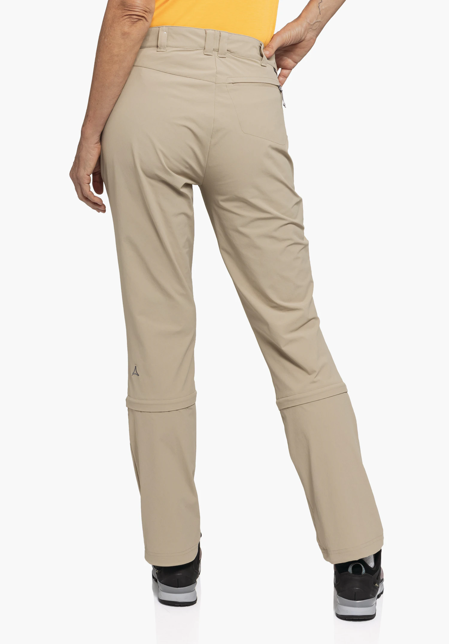 Pants Engadin1 Zip Off getragen in der Rückenansicht von einem Model. Man sieht die beige Hose mit charakteristischen Zip-Off-Reißverschlüssen über den Knien, Gürtelschlaufen und einer Gesäßtasche mit Reißverschluss auf der rechten Seite. Das Schöffel-Logo ist auf der Rückseite des linken Beins sichtbar. Das Model steht vor einem neutralen Hintergrund und kombiniert die Hose mit einem orangefarbenen Oberteil und dunklen Wanderschuhen.