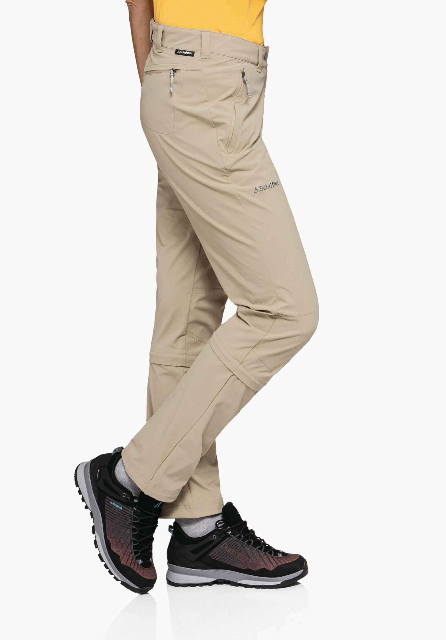 Pants Engadin1 Zip Off in Beige an einem Model, das seitlich steht und die Zip-Off-Reißverschlüsse sowie das Schöffel-Logo auf dem rechten Oberschenkel zeigt, präsentiert vor einem neutralen Hintergrund.