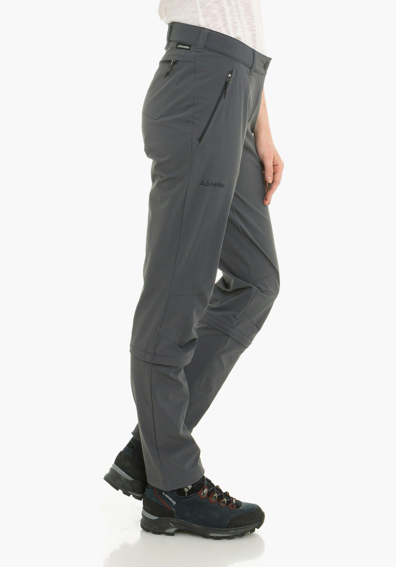 Pants Engadin1 Zip Off dark grey outdoor trousers with Schöffel logo and lettering "Schöffel", worn side-on; person stands side-on with one leg slightly forward, white background; dark blue hiking boots.