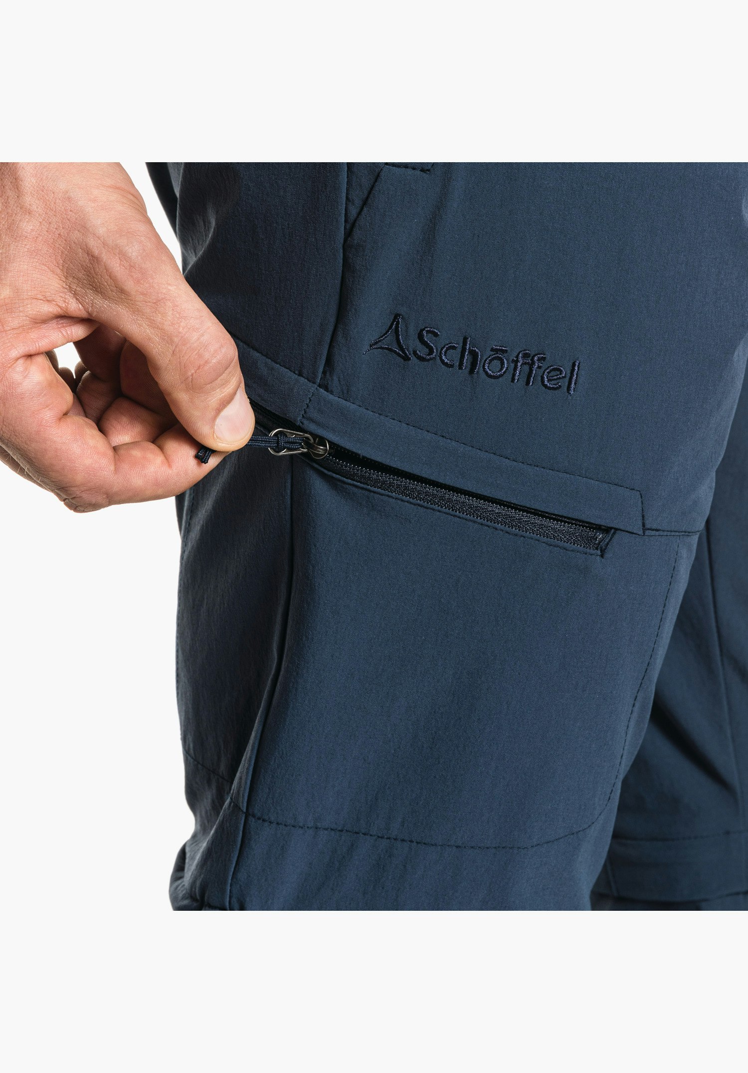Pants Koper1 Zip Off zeigt den Reißverschluss der Oberschenkeltasche auf dem blauen Stoff sowie das gestickte Schöffel-Logo. Nahaufnahme vor weißem/neutralem Hintergrund.