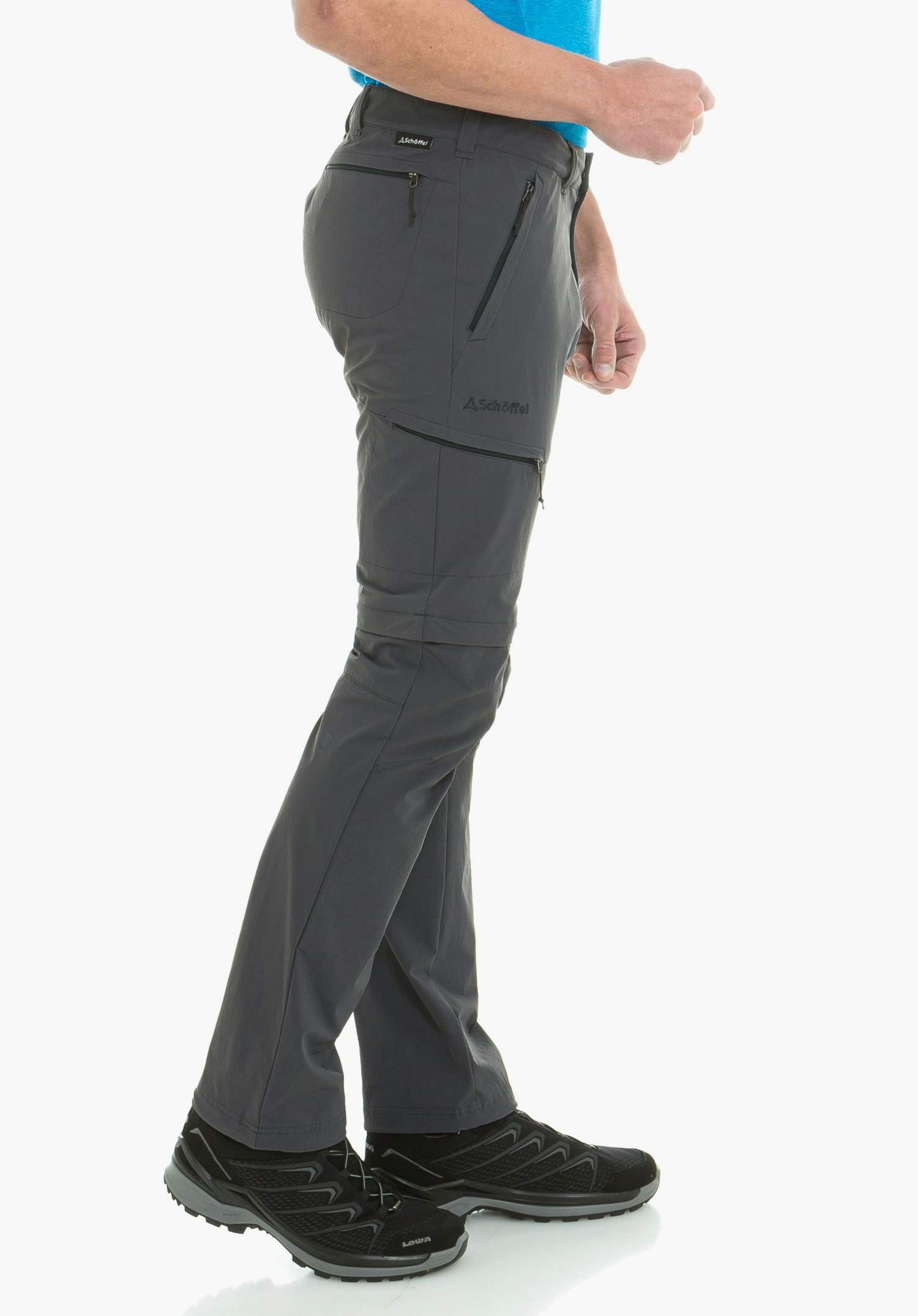 Pants Koper1 Zip Off pantalon outdoor gris foncé porté par une personne debout avec t-shirt bleu clair et chaussures de randonnée noires; poches zippées latérales, logo Schöffel, textes: "Schöffel", "LOWA".