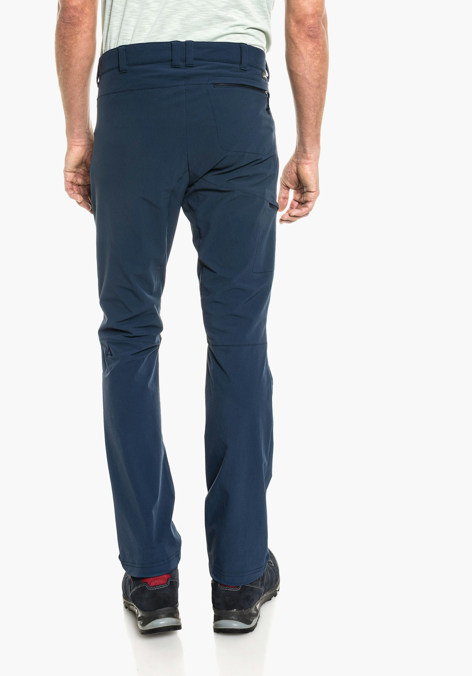 Pants Koper1 pantalon bleu foncé, porté de dos par une personne debout sur fond blanc; poche arrière zippée, logo Schöffel à la taille, bord de T-shirt vert clair visible, chaussures bleu foncé avec accent rouge au talon.