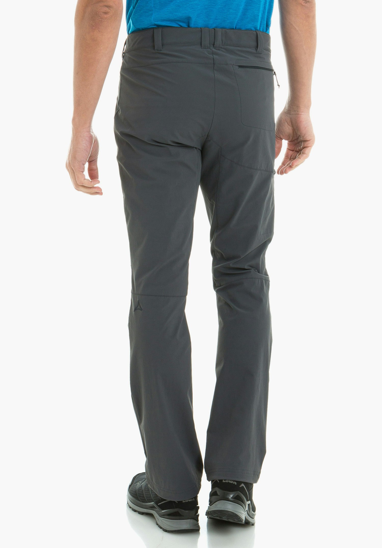 Pants Koper1 pantalon outdoor gris foncé porté par un homme, debout dos à l’objectif; logo Schöffel sur la jambe gauche; T-shirt bleu clair partiellement visible; chaussures de randonnée noires avec inscription « LOWA »; fond studio blanc.