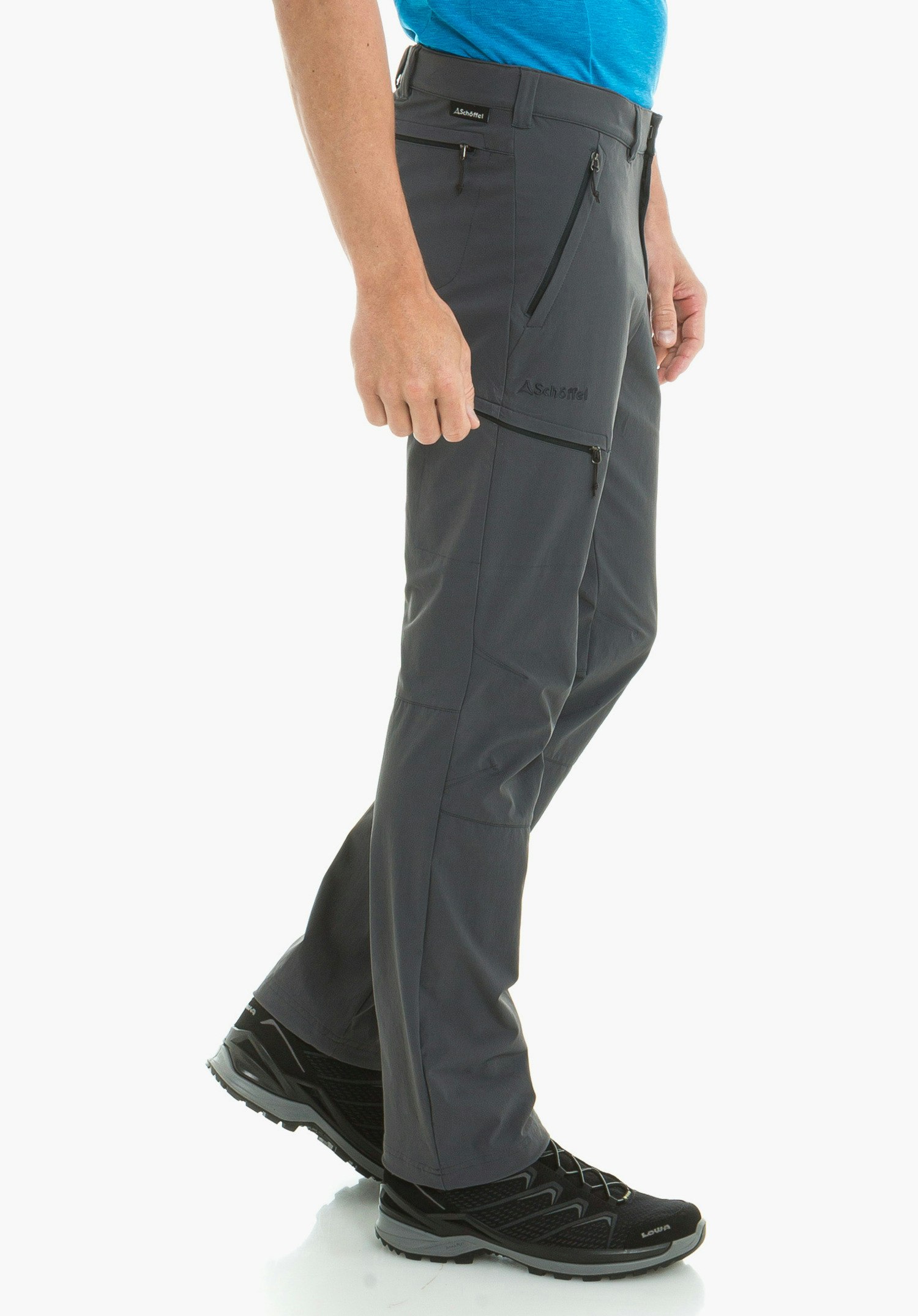 Pants Koper1 pantalon de trekking gris foncé, porté; la personne avance légèrement devant un fond clair uni; t-shirt bleu clair, chaussures de randonnée noires. Logo Schöffel visible. Textes: Schöffel; LOWA.