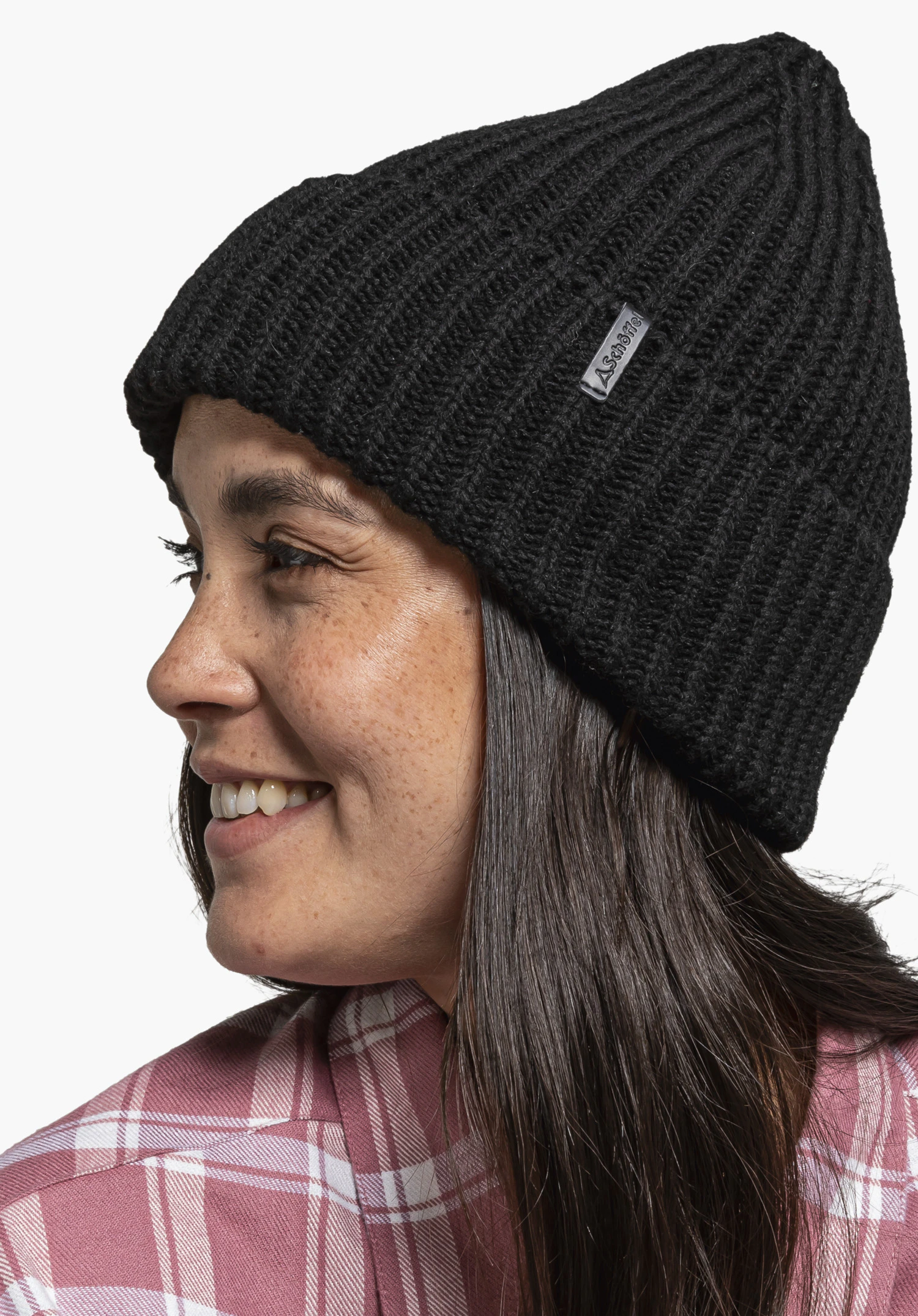 Knitted Hat Medford schwarze Strickmütze, von einer Frau lächelnd zur Seite getragen; neutraler Hintergrund, sie trägt rosa kariertes Hemd und hat dunkelbraune Haare. Auf Mütze Schöffel-Logo, Schrift "Schöffel".