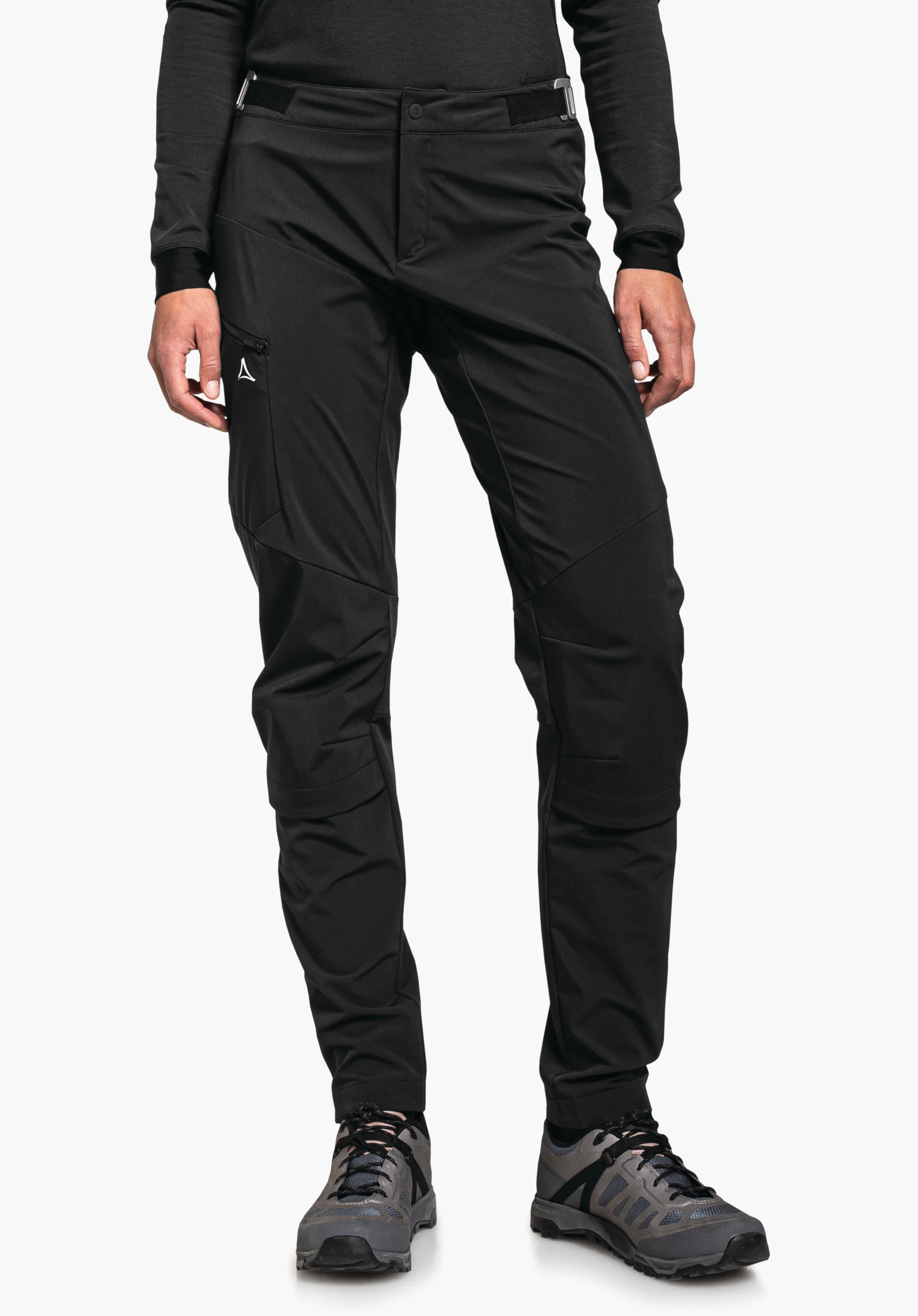 Hybrid Pants Tree Run L pantalon outdoor noir avec logo Schöffel sur la poche de cuisse, porté par une personne debout; bras le long du corps, chaussures de randonnée grises; fond blanc.
