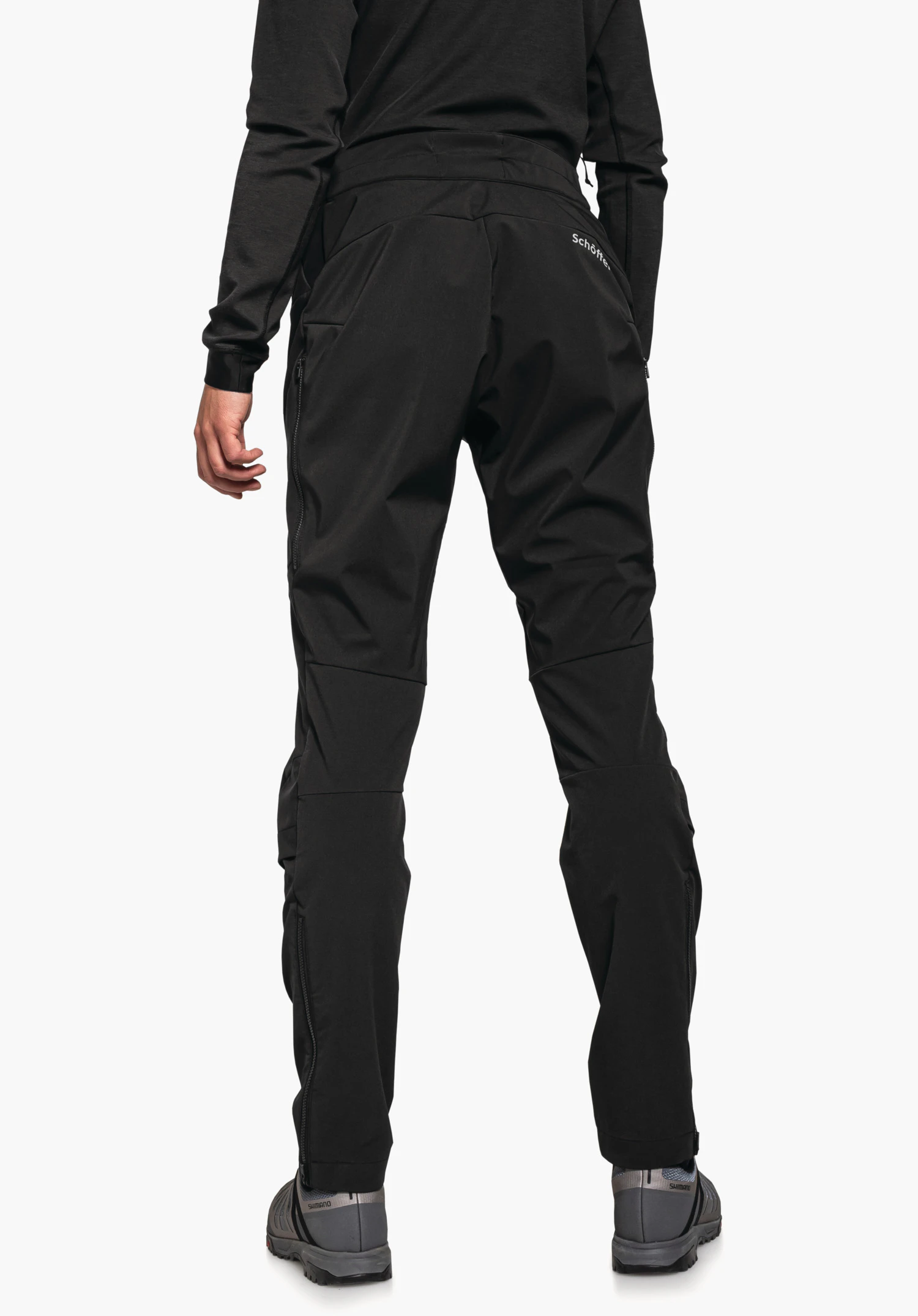 Hybrid Pants Tree Run L pantalon outdoor noir, porté par une personne debout vue arrière sur fond blanc; texte « Schöffel » sur la poche arrière droite, chaussures avec texte « SHIMANO »; haut noir, chaussures gris foncé.