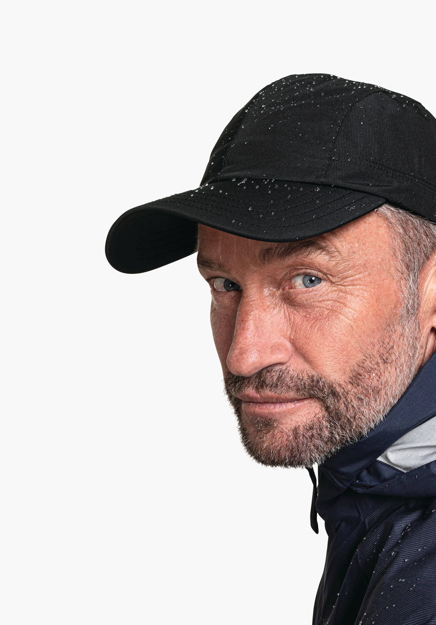 Rain Cap3 in Schwarz: Ein männliches Model präsentiert die Cap in einer seitlichen Nahaufnahme vor einem neutralen Hintergrund. Die Kappe ist mit Wassertropfen bedeckt, was die Wetterfestigkeit unterstreicht. Das Schöffel-Logo ist diskret auf der Seite platziert.