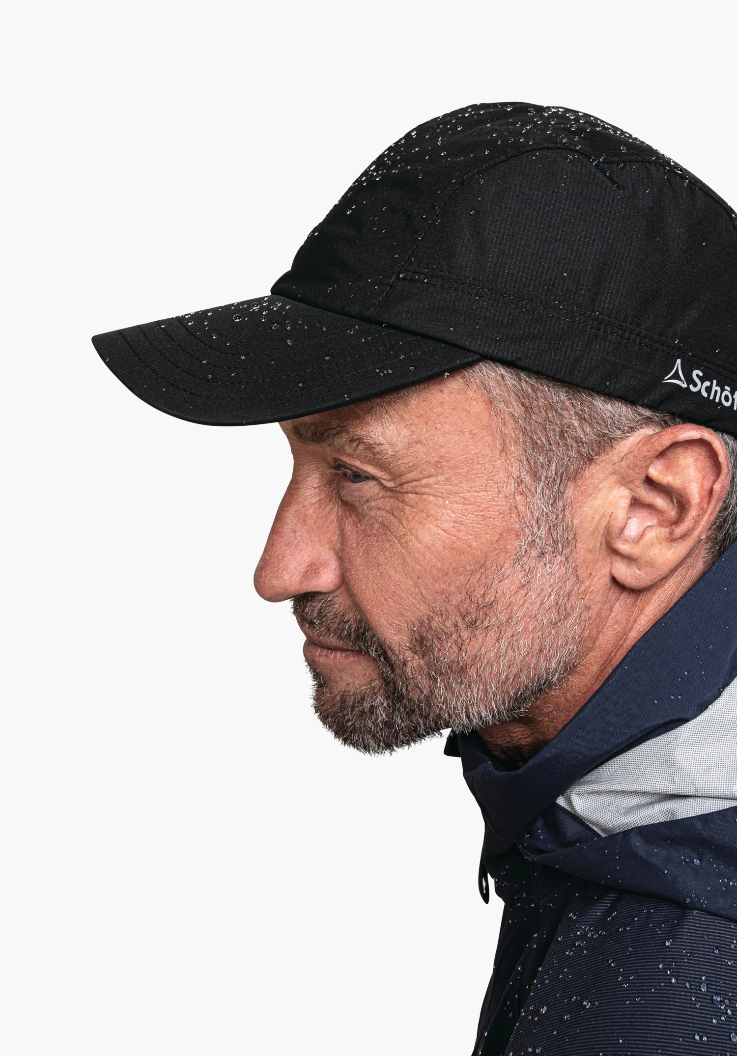 Rain Cap3 getragen von einem Model in Seitenansicht. Die Kappe in Schwarz ist mit Wassertropfen bedeckt und an der Seite ist das Schöffel-Logo deutlich sichtbar. Das Model steht vor einem neutralen Hintergrund.