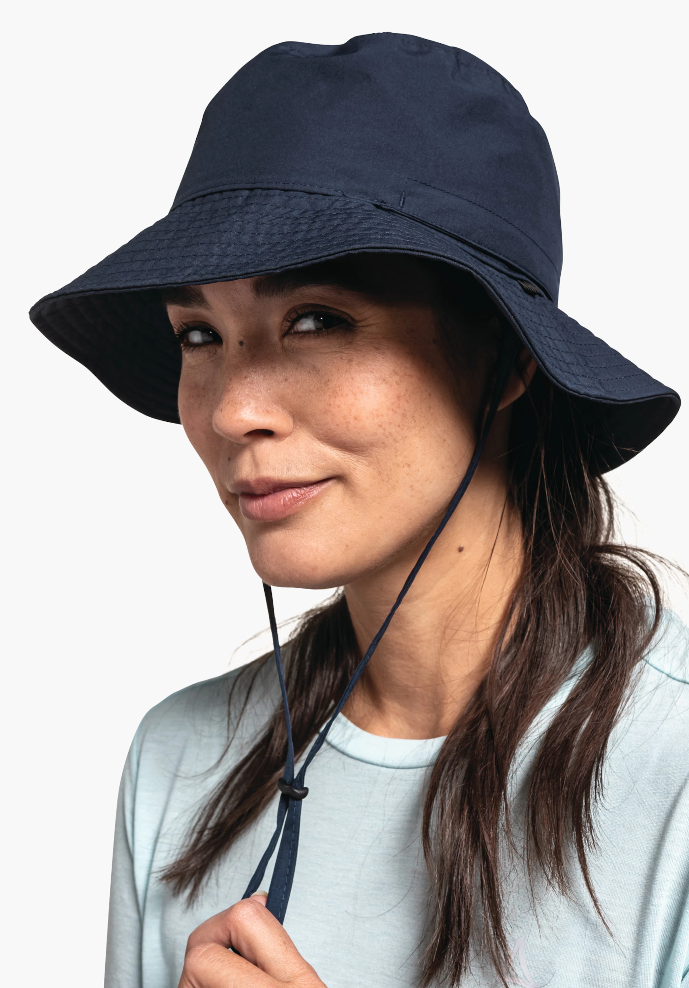 Rain Hat4 in Blau wird hier von einer Person getragen, die frontal mit einer leichten Kopfdrehung in die Kamera blickt. Der Hut mit praktischem Kinnband sitzt perfekt vor einem neutralen Hintergrund, wobei das dezente Schöffel-Logo an der Seite erkennbar ist.