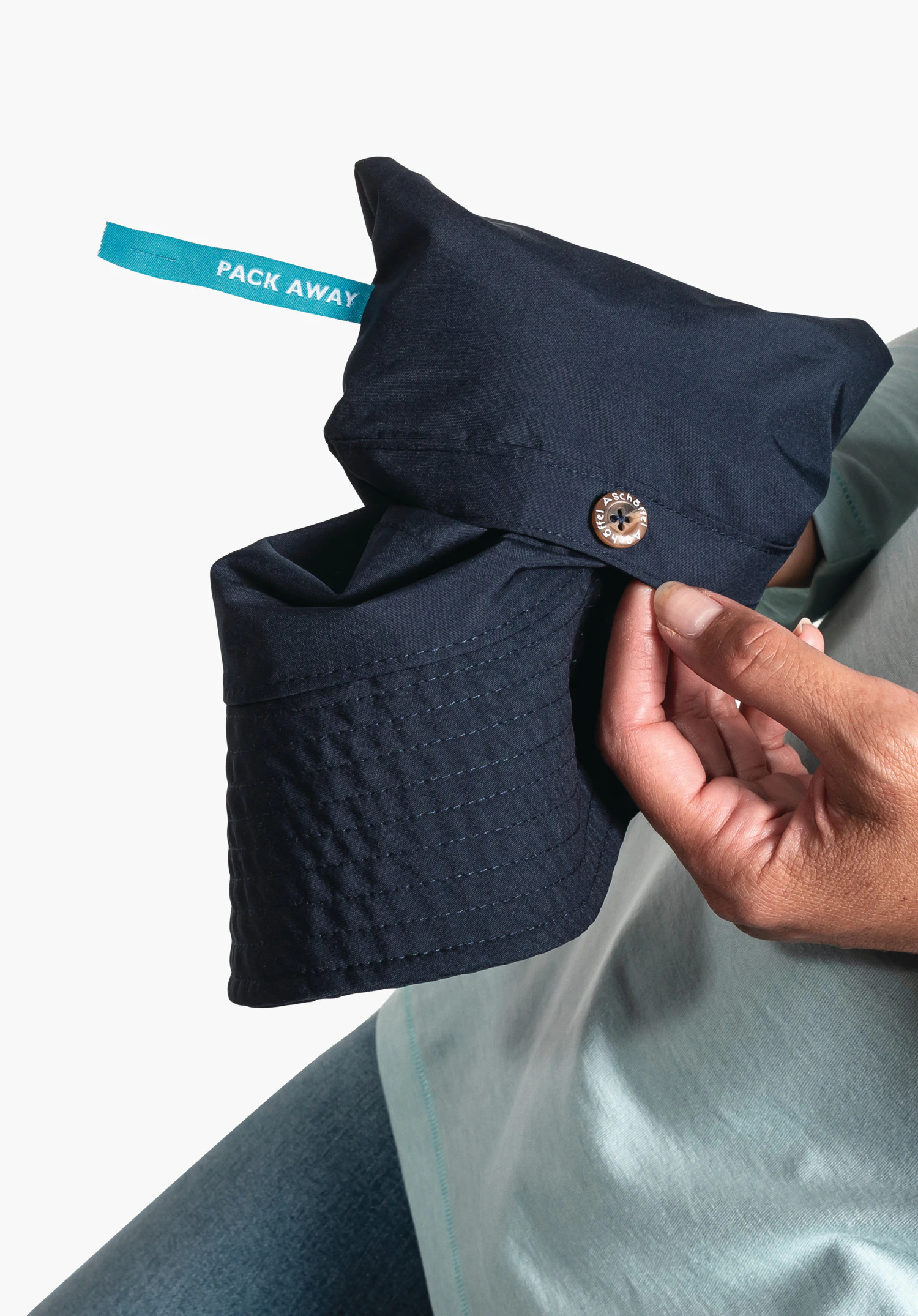 Rain Hat4 in Blau, das Detail zeigt das hellblaue "PACK AWAY"-Label und den Knopf mit Schöffel-Logo am zusammengefalteten Stoff in den Händen einer Person. Nahaufnahme vor weißem Hintergrund.