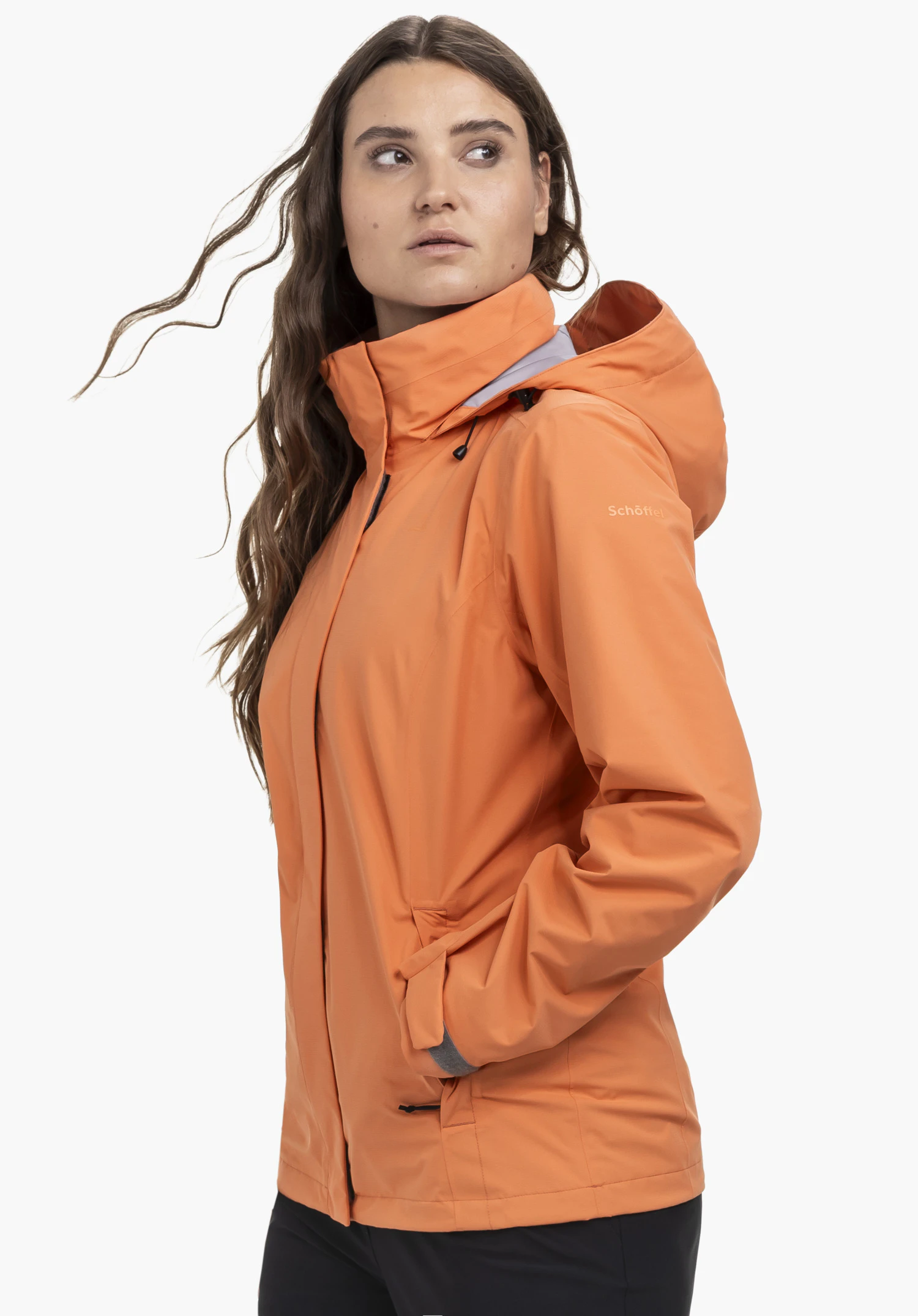 Jacket Gmund L worn by a model standing sideways with her hand in the side pocket. The orange hooded jacket clearly shows the Schöffel-Logo on the left sleeve, photographed against a neutral background.
