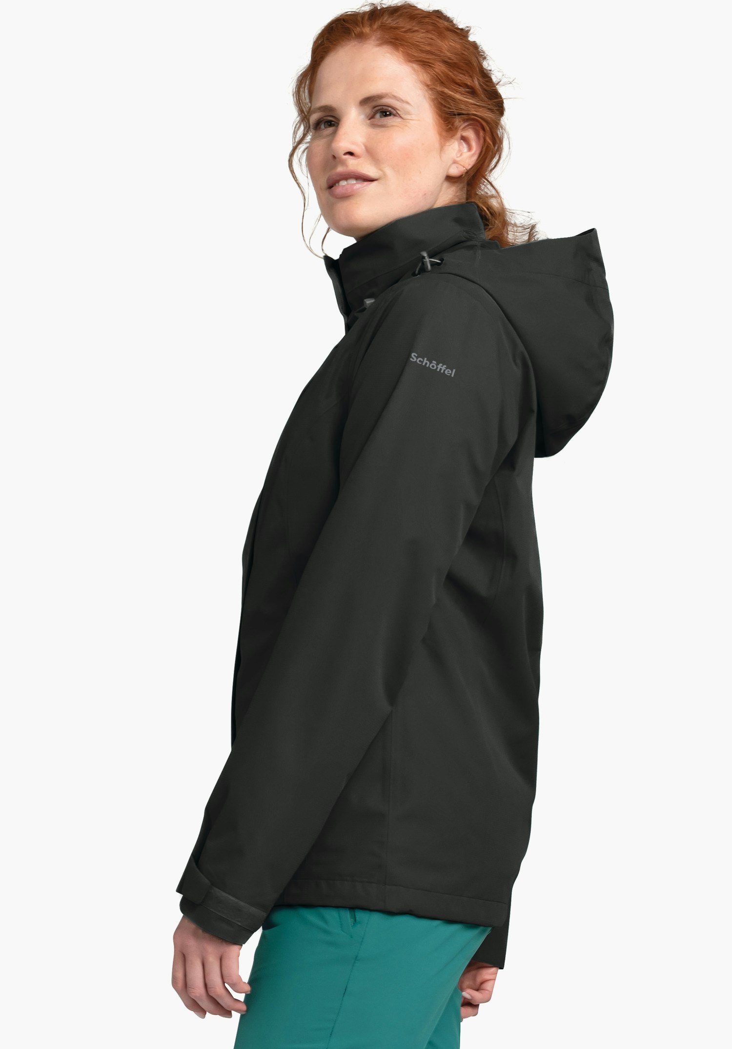 Jacket Gmund L Dunkelgraue Regenjacke, von einer Frau seitlich getragen, mit hochgestellter Kapuze; Frau blickt leicht nach oben. Kontext: heller neutraler Hintergrund. Ärmeltext: "Schöffel".