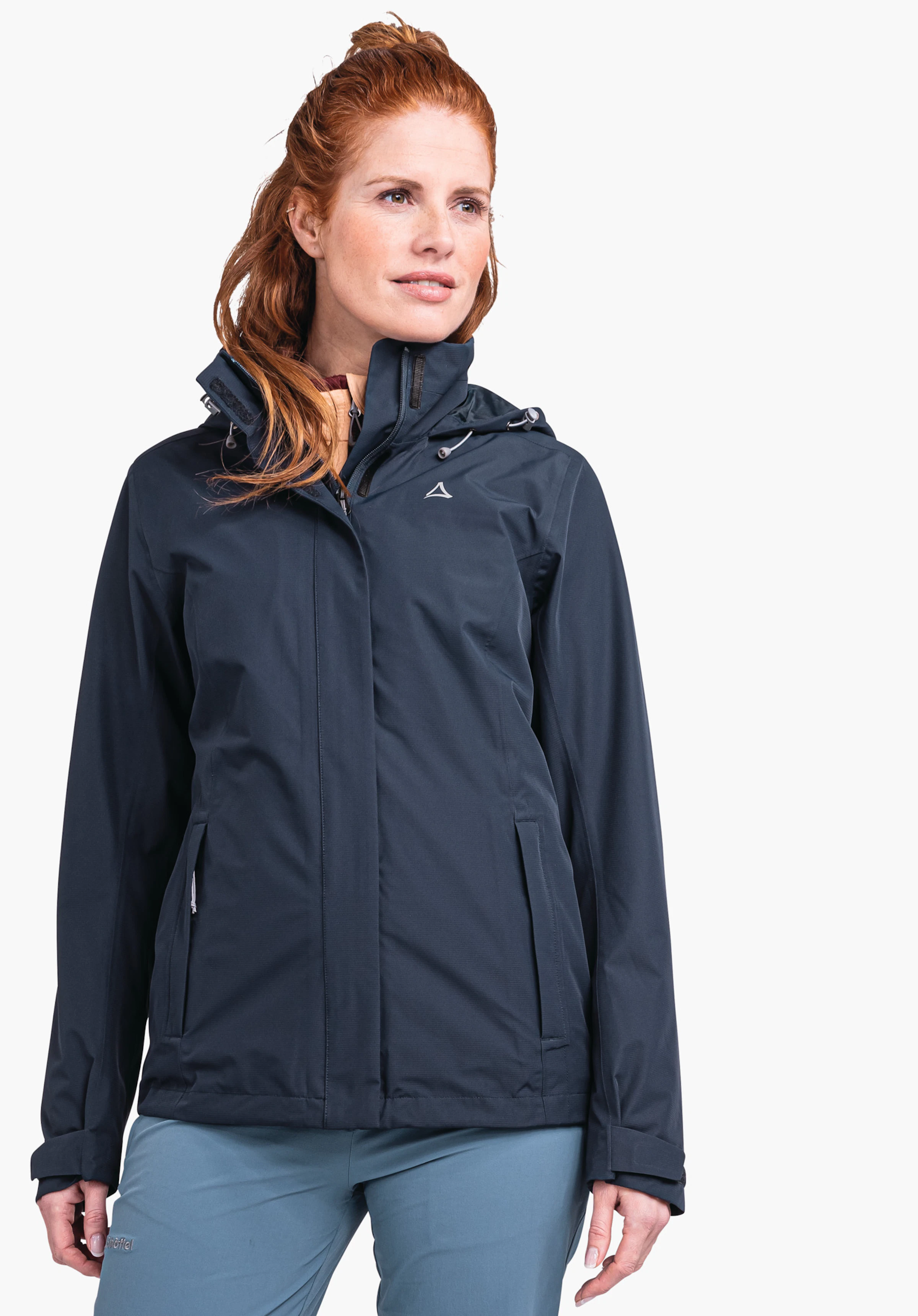 Jacket Gmund L Dunkelblaue Jacke mit Schöffel-Logo, getragen von Frau mit roten Haaren; sie steht leicht seitlich und blickt nach rechts vor weißem Studiohintergrund, hellblaue Hose sichtbar.