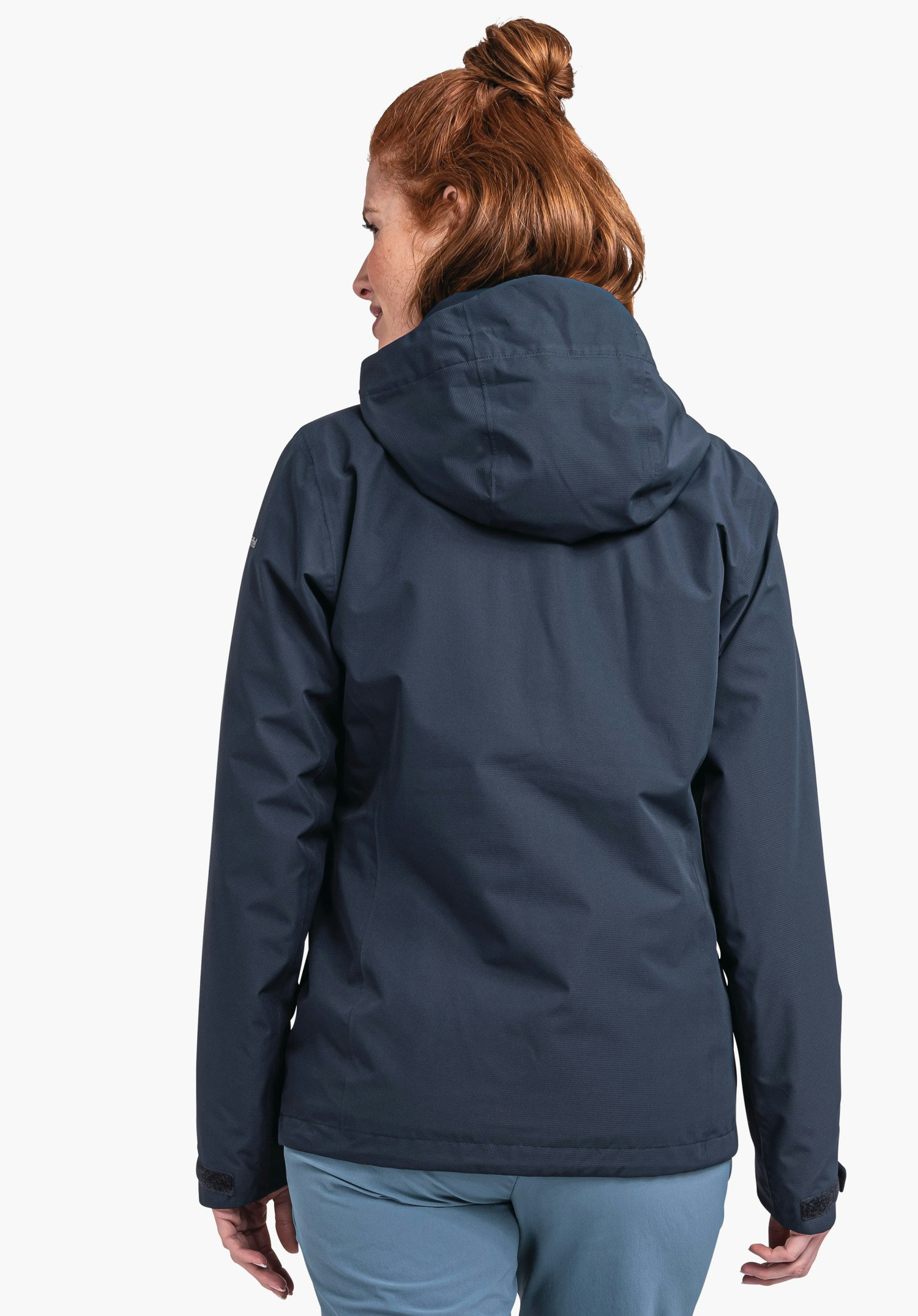 Jacket Gmund L Frau in dunkelblauer Regenjacke steht mit dem Rücken zur Kamera, dreht leicht den Kopf nach links; trägt hellblaue Hose; rote Haare zu einem Dutt; weißer Hintergrund.