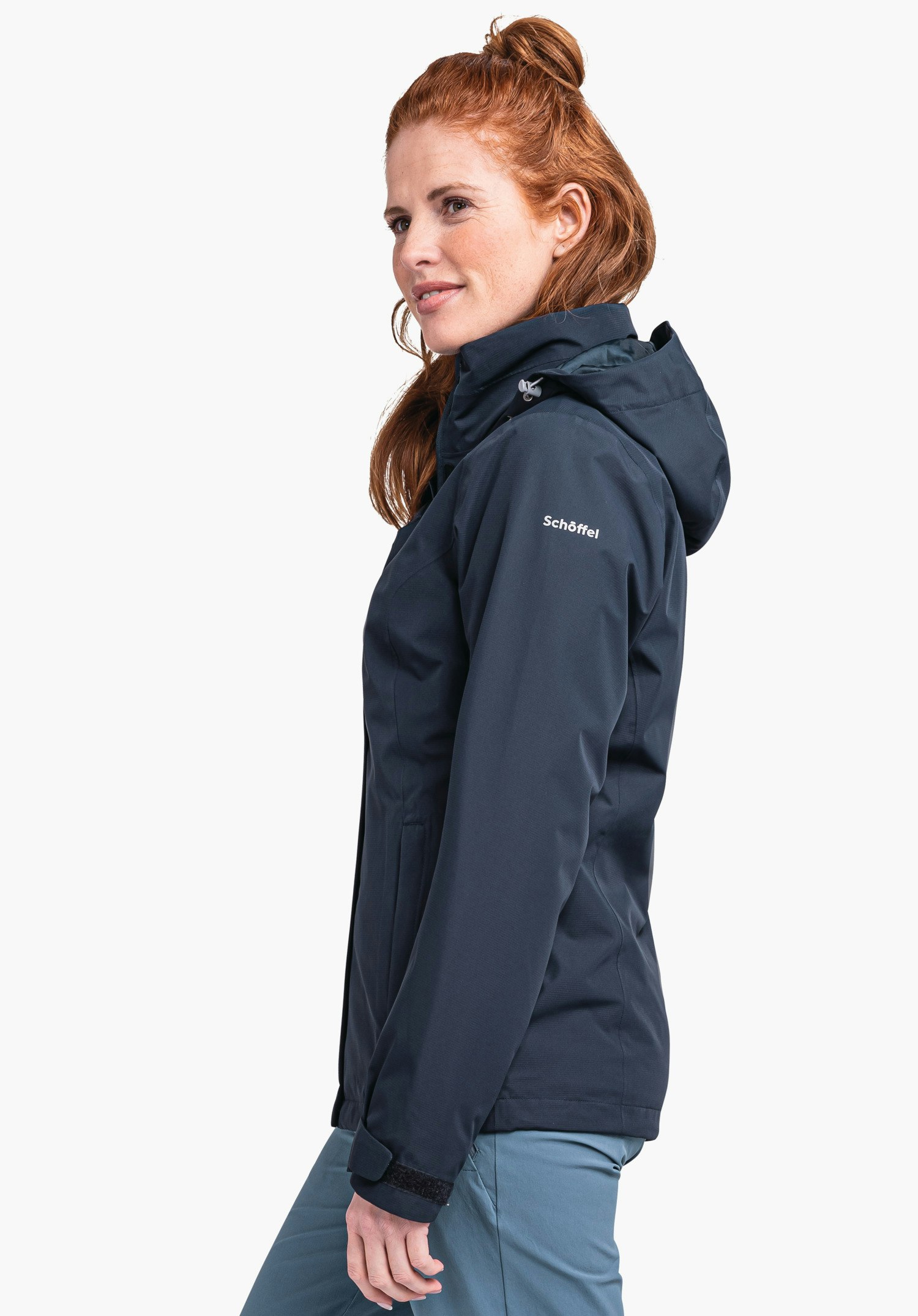 Jacket Gmund L dunkel blau Jacke, seitlich getragen von einer Frau, sie lächelt, Kapuze auf dem Rücken; hell blau Hose; rotes Haar; weißer Studiohintergrund. Text auf Ärmel: "Schöffel".