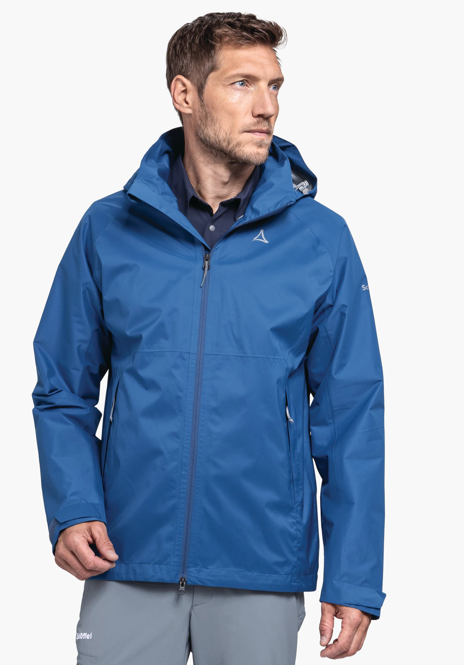 Jacket Easy XT M. Le mannequin se tient de face dans la veste bleue et regarde légèrement sur le côté. Le Schöffel-Logo est bien visible sur la poitrine gauche, présenté devant un fond neutre.
