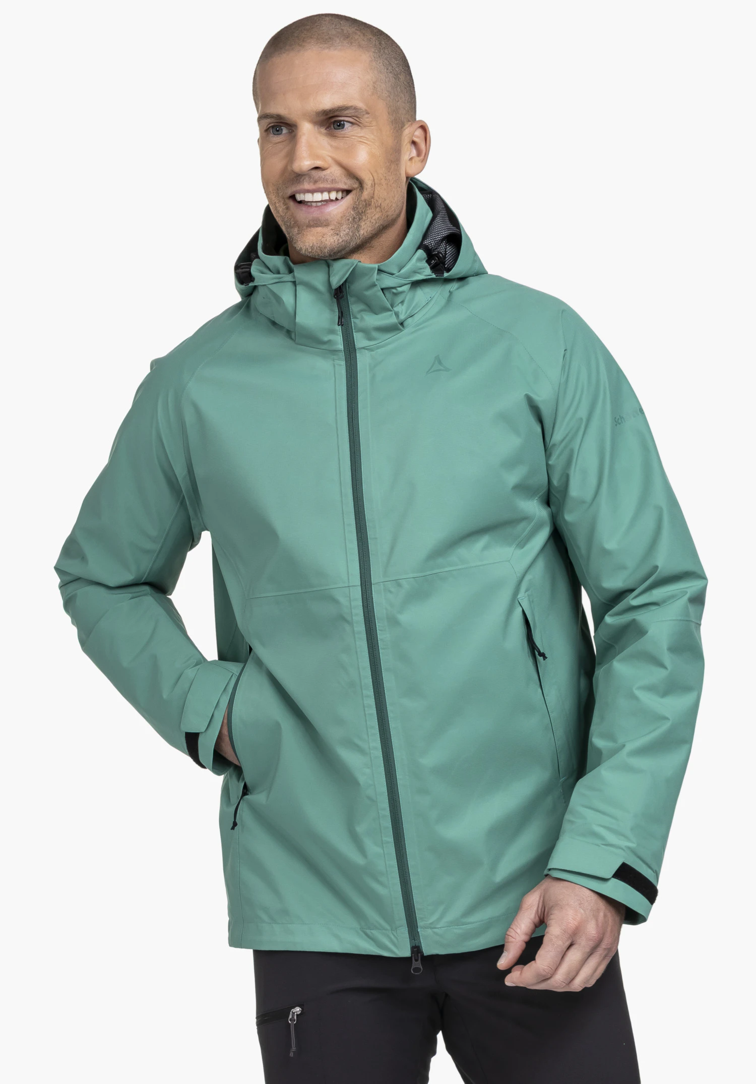 Jacket Easy XT M gedragen door een lachend model dat in een lichte zijaanzicht staat. De groene jas heeft een vaste capuchon, een gesloten ritssluiting aan de voorkant en het Schöffel-logo op de linkerborst, waarbij één hand ontspannen in de zijzak rust, gefotografeerd tegen een neutrale achtergrond.