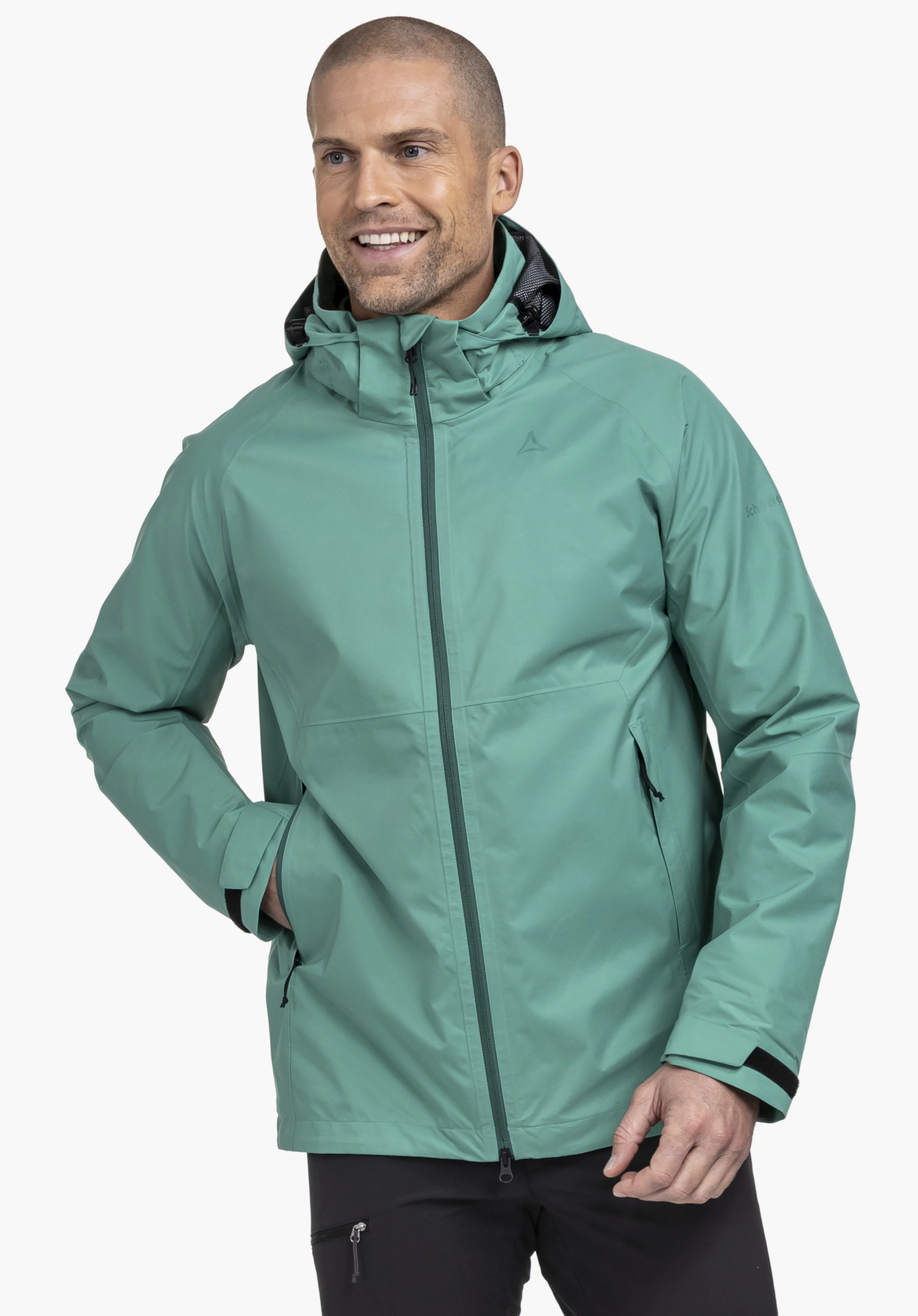 Jacket Easy XT M gedragen door een lachend model dat in een lichte zijaanzicht staat. De groene jas heeft een vaste capuchon, een gesloten ritssluiting aan de voorkant en het Schöffel-logo op de linkerborst, waarbij één hand ontspannen in de zijzak rust, gefotografeerd tegen een neutrale achtergrond.